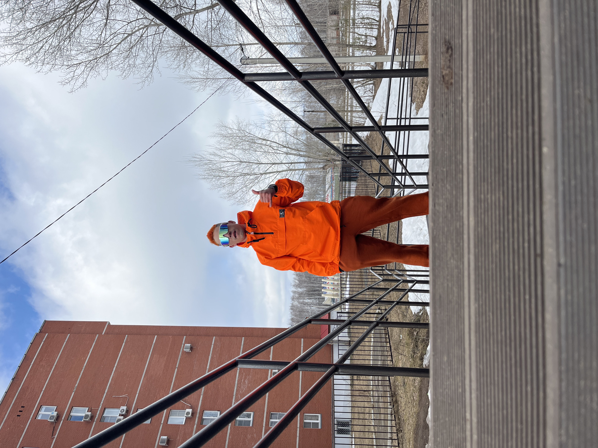 A man in an orange jacket and pants, wearing mirrored goggles, stands on a platform with metal railings in Chaikovsky, Russia, giving a thumbs up. The sky is overcast, and there are bare trees and a brick building in the background. Snow patches are visible on the ground.