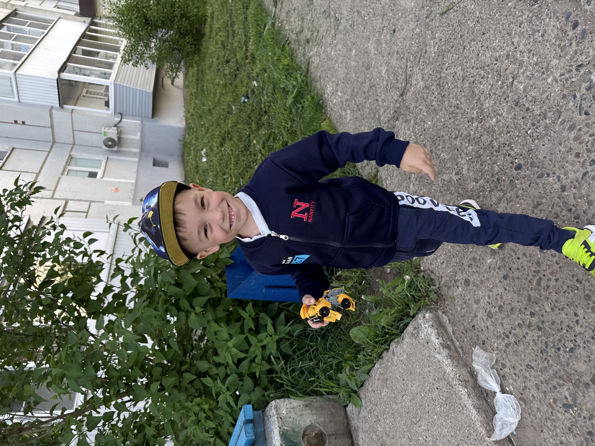 The image portrays a young boy, likely a toddler or preschooler, standing outdoors on a concrete path in Chaikovsky, Russia. He is positioned slightly off-center and looks directly at the camera with a wide, joyful smile, his head tilted. The boy is dressed in a dark blue zip-up hoodie or jacket with a white collar, dark blue sweatpants featuring white lettering down the side, and vibrant yellow-green sneakers. A baseball cap with a yellow brim and a dark, patterned crown completes his playful attire.
In his right hand, the boy proudly holds a yellow toy vehicle, resembling a transformer or a construction truck. His cheerful expression suggests he is happy and engaged in play.
The background presents a mix of urban and natural elements. Lush green bushes and trees flank the left side and foreground, indicating a grassy area or park. Behind the boy, a multi-story apartment building with a facade of windows and balconies is visible. An air conditioning unit is attached to the building's exterior. The ground beneath the boy is a textured concrete path, and a small, crumpled white plastic bag lies on it in the lower right corner. A blue wooden structure, possibly a planter or low fence, is partly visible near his feet.
Visible text on the boy's jacket includes a prominent red letter "N" with "NAVVITY" written underneath it, and another partly obscured word, "NANNTITY." The white lettering on his pants is not clearly decipherable. The scene takes place during the daytime under soft, even lighting, indicative of pleasant weather. The architectural style of the background building is characteristic of residential constructions found in post-Soviet regions.