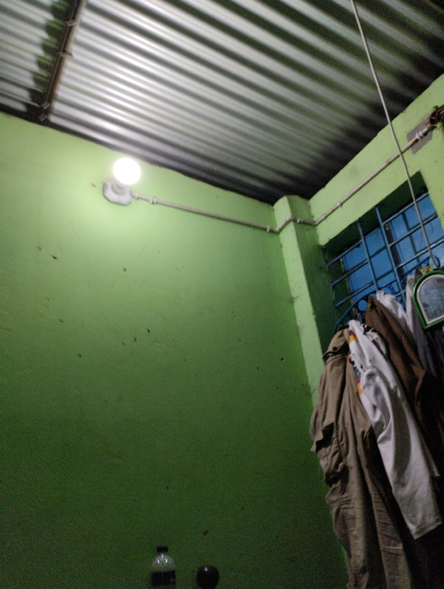 An indoor scene in a dimly lit room in Dhaka, Bangladesh. The room features light green walls and a corrugated metal ceiling. A single light bulb illuminates the area, casting a glow on the wall. On the right side of the image, clothes are hanging on a rack in front of a barred window. A bottle of water and a dark spherical object are visible on the floor in the lower part of the frame.