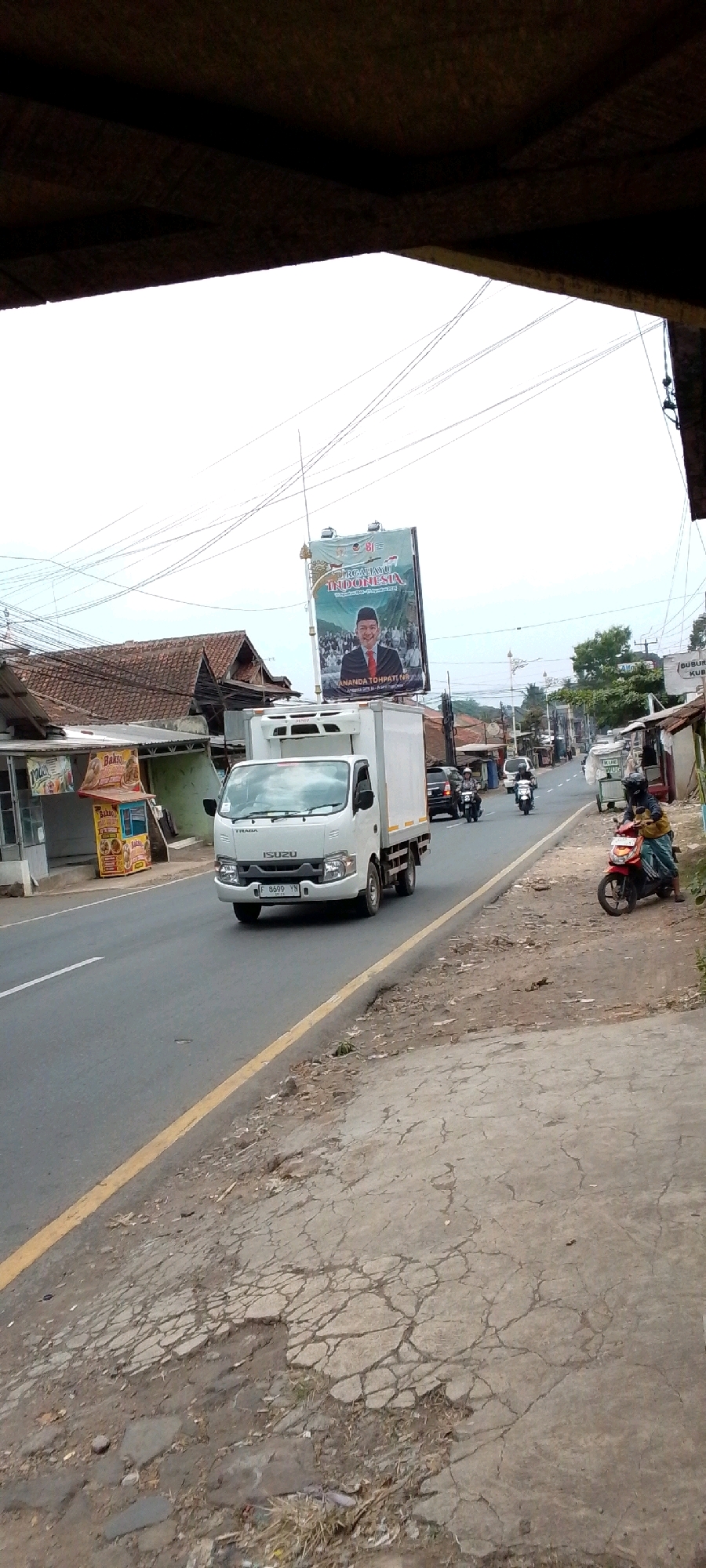 The image captures a street scene in Cilaku, Indonesia, under an overcast sky. A white Isuzu truck is in motion on a paved road, its license plate reading "F 8699 YN". To the left of the road, a small food stall with yellow and red signage is visible. Further left are buildings with terracotta tile roofs. A large billboard featuring a man in a suit and the text "PERGATUHUI INDONESIA" and "ANANDA TOHPATI" is prominently displayed. Several motorcycles are also on the road, with two riders visible. The right side of the road features a cracked concrete surface with sparse vegetation. The scene is framed from below by a dark, textured overhang, likely part of a building or structure.