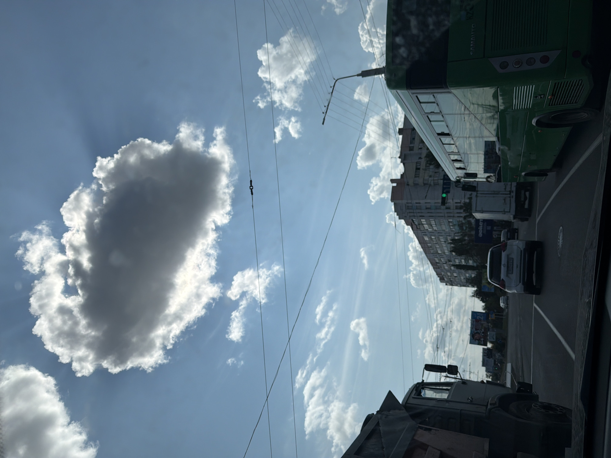 The image depicts a city street scene in Pavlodar, Kazakhstan, under a partly cloudy sky. The perspective is from inside a vehicle, looking forward. In the foreground, the edge of a dashboard or car interior is visible.
The primary focus of the scene is a street intersection with multiple vehicles. To the left, a large, grey dump truck is partially visible. In the center lane, a white SUV is driving away from the camera, and to its right, a long, green bus with "SAMAUTO" branding on the back is also moving forward. Other vehicles, including what appears to be a white truck, are visible in the background.
The environment includes multi-story buildings lining the street, power lines crisscrossing the sky, and traffic signals. One traffic light, showing green, is visible near the bus. Billboards are also present in the distance. The clouds in the sky are dramatic, with a large, dark cloud with sunbeams radiating from behind it dominating the upper portion of the frame. The overall impression is a typical urban scene on a day with dynamic weather. There are no visible people in the image.