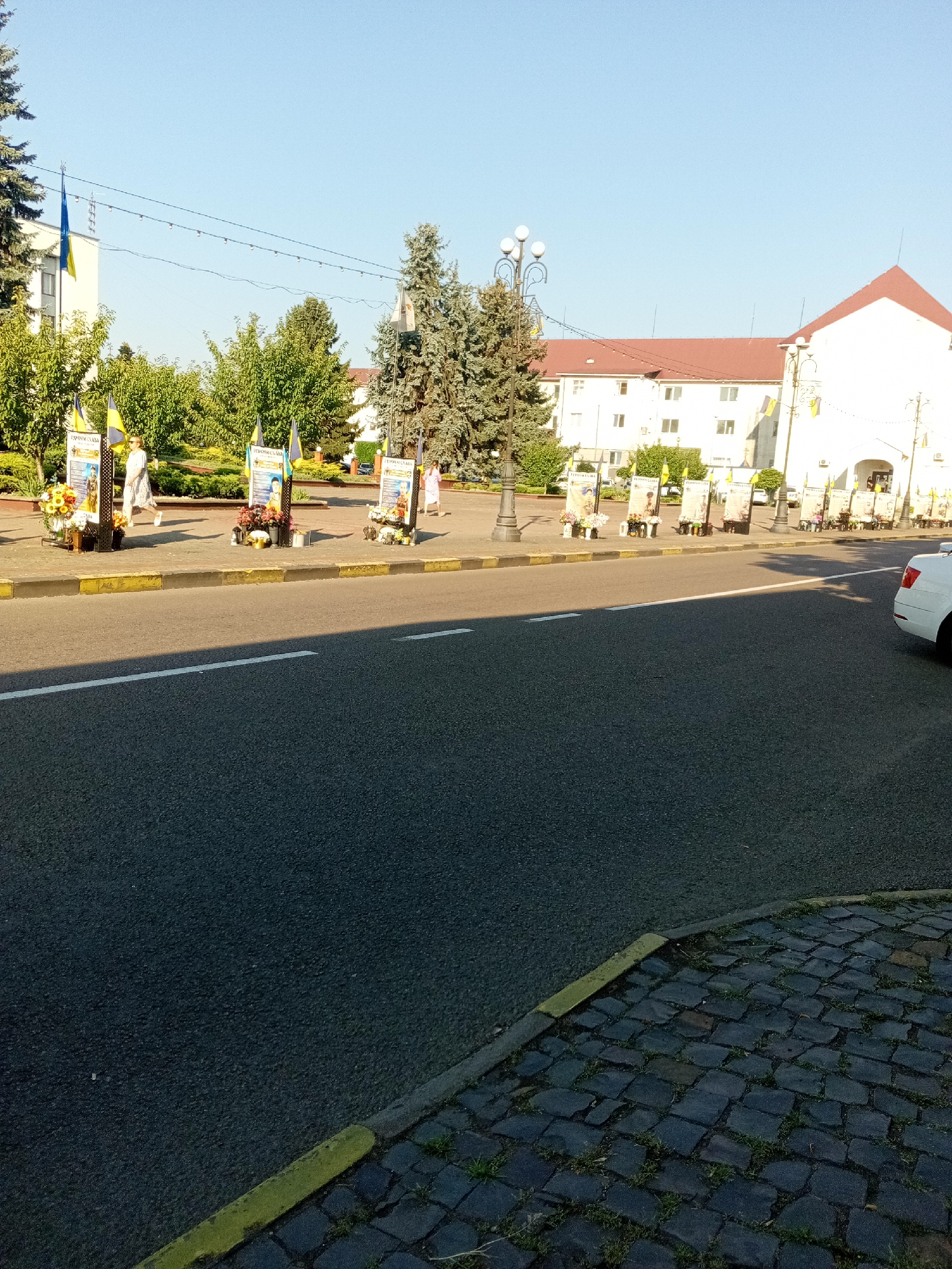 The image depicts a scene in Tiachiv, Ukraine, likely a public square or street. Several memorial displays are set up along a sidewalk, featuring portraits and floral arrangements. Ukrainian flags are prominently displayed, indicating a patriotic or commemorative event. A white car is partially visible on the right side of the road, and people are walking in the background. The environment appears to be a sunny day with clear blue skies, set against a backdrop of buildings and trees. The foreground consists of a paved road with a yellow curb.