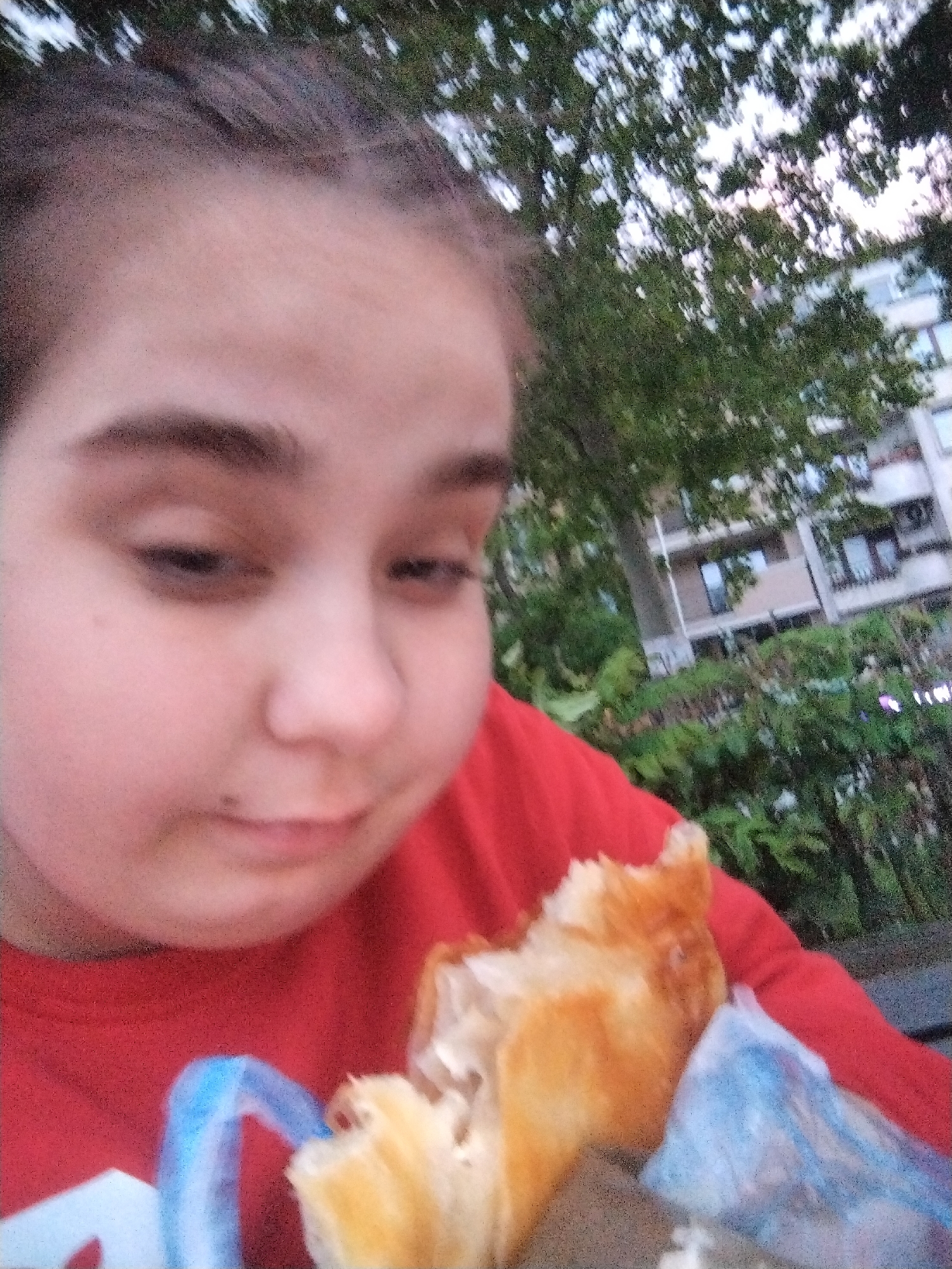A young person is holding a pastry outside in what appears to be the city of Plovdiv, Bulgaria. The person has fair skin, dark eyebrows, and their hair is pulled back. They are wearing a red shirt. In their hand, they hold a partially eaten pastry with a flaky, golden-brown crust. Part of a blue plastic bag is visible next to the pastry. Behind the person are green trees and a building with windows, suggesting an urban park setting.