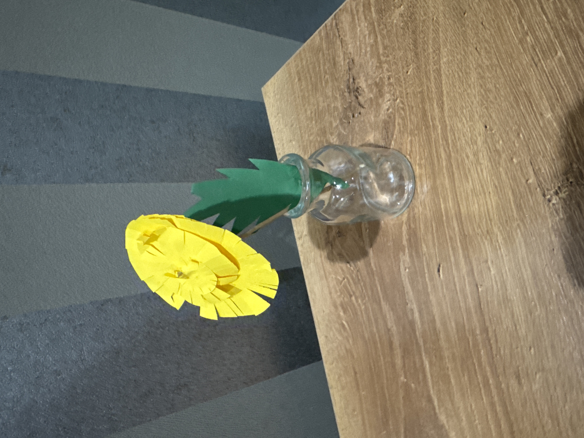 A handmade yellow paper flower with a green paper leaf stands in a small clear glass vase on a wooden surface. The background is a textured dark gray wall with vertical lighter gray stripes. The wooden surface exhibits a prominent grain pattern and appears to be a tabletop or shelf.