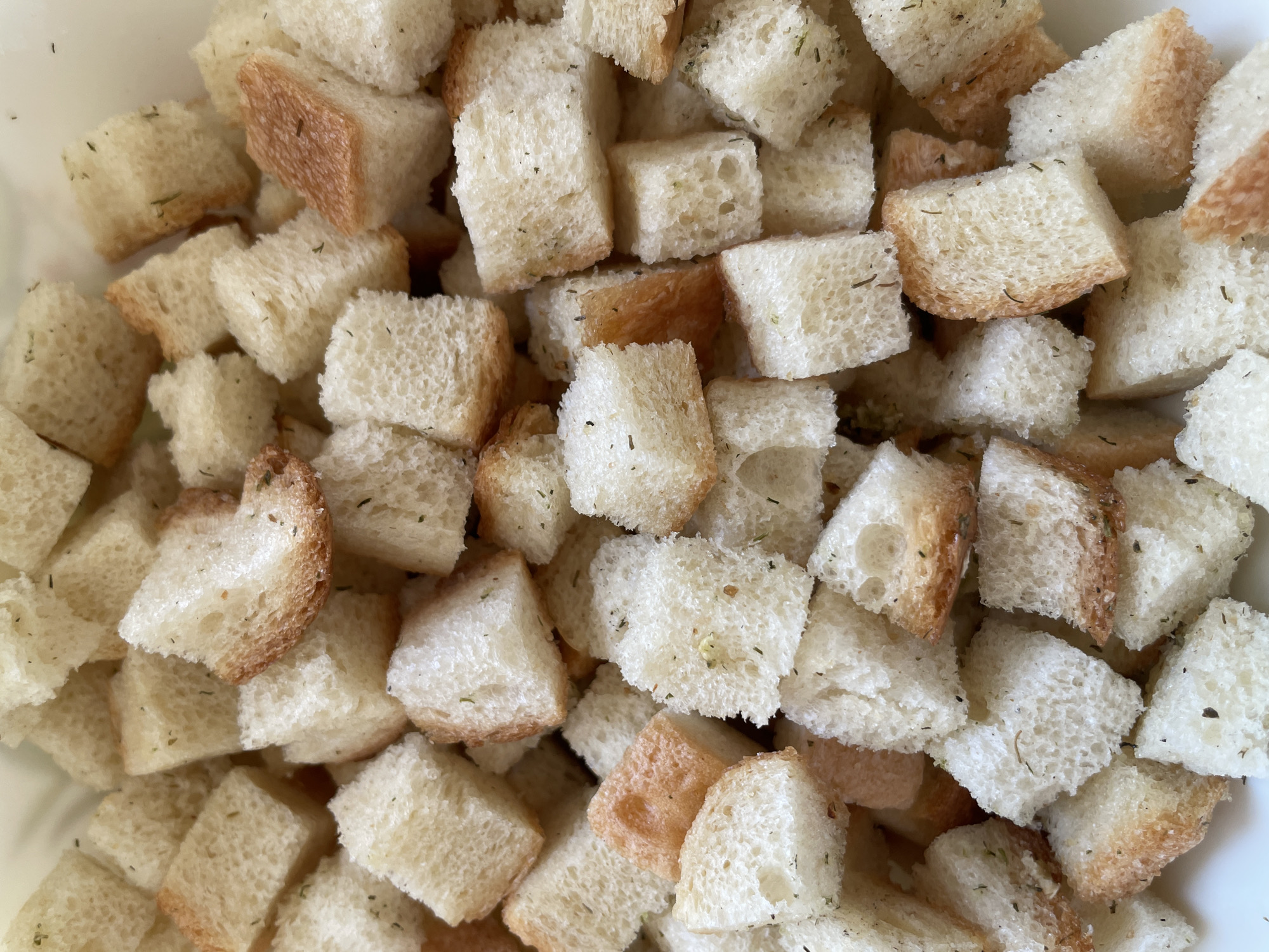 The image shows a close-up of a pile of croutons. The croutons are mostly cube-shaped, with a light tan color on some edges and white in the center. Small green flecks, possibly herbs, are scattered on the croutons. The croutons appear to be contained in a white bowl or container. The location is unknown.