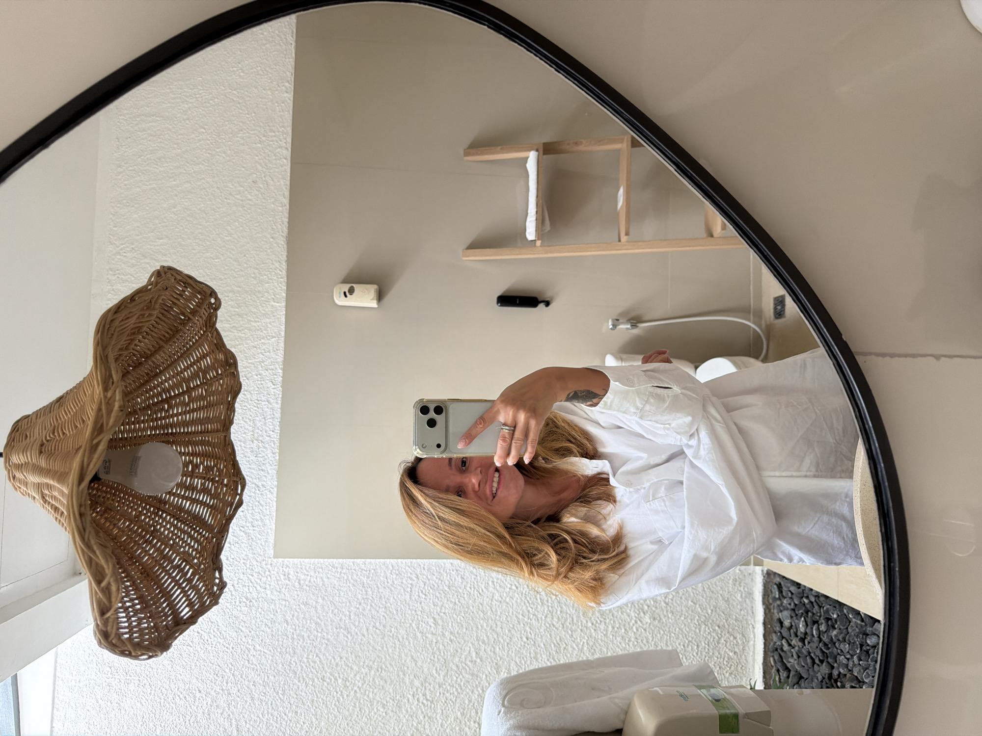 A woman with auburn hair, wearing a white oversized button-down shirt, takes a selfie in a mirror. She is smiling and holding a smartphone in her right hand. A ring is visible on her left ring finger. Behind her, reflected in the mirror, is a bathroom with neutral-colored walls. A woven pendant light hangs from the ceiling. To the right, a wooden ladder shelf holds a folded white towel. A toilet and a wall-mounted dispenser are also visible. The bathroom floor has small black stones. The scene is likely in a hotel or guesthouse in Bali, Indonesia, given the aesthetic and context.