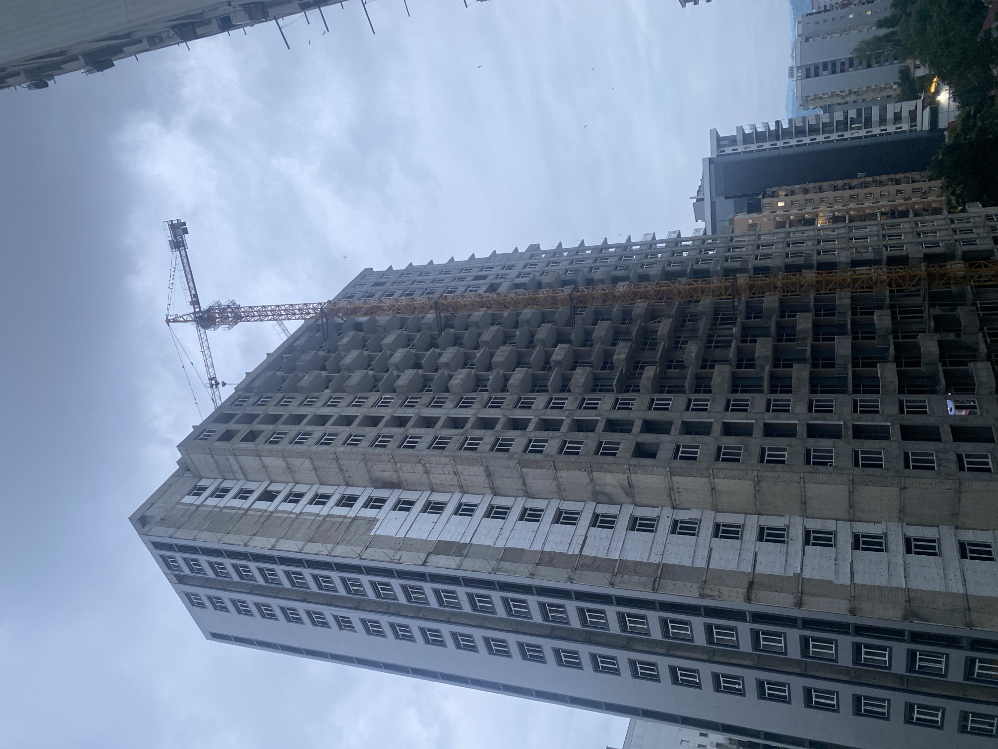 A low-angle, vertical outdoor shot shows a massive, unfinished apartment building under construction in the city. The building is made of concrete and has many floors. A yellow construction crane is attached to the side of the building. The sky is overcast and gray. There are other buildings visible in the background, as well as trees. The scene appears to be a typical urban construction site on a cloudy day.