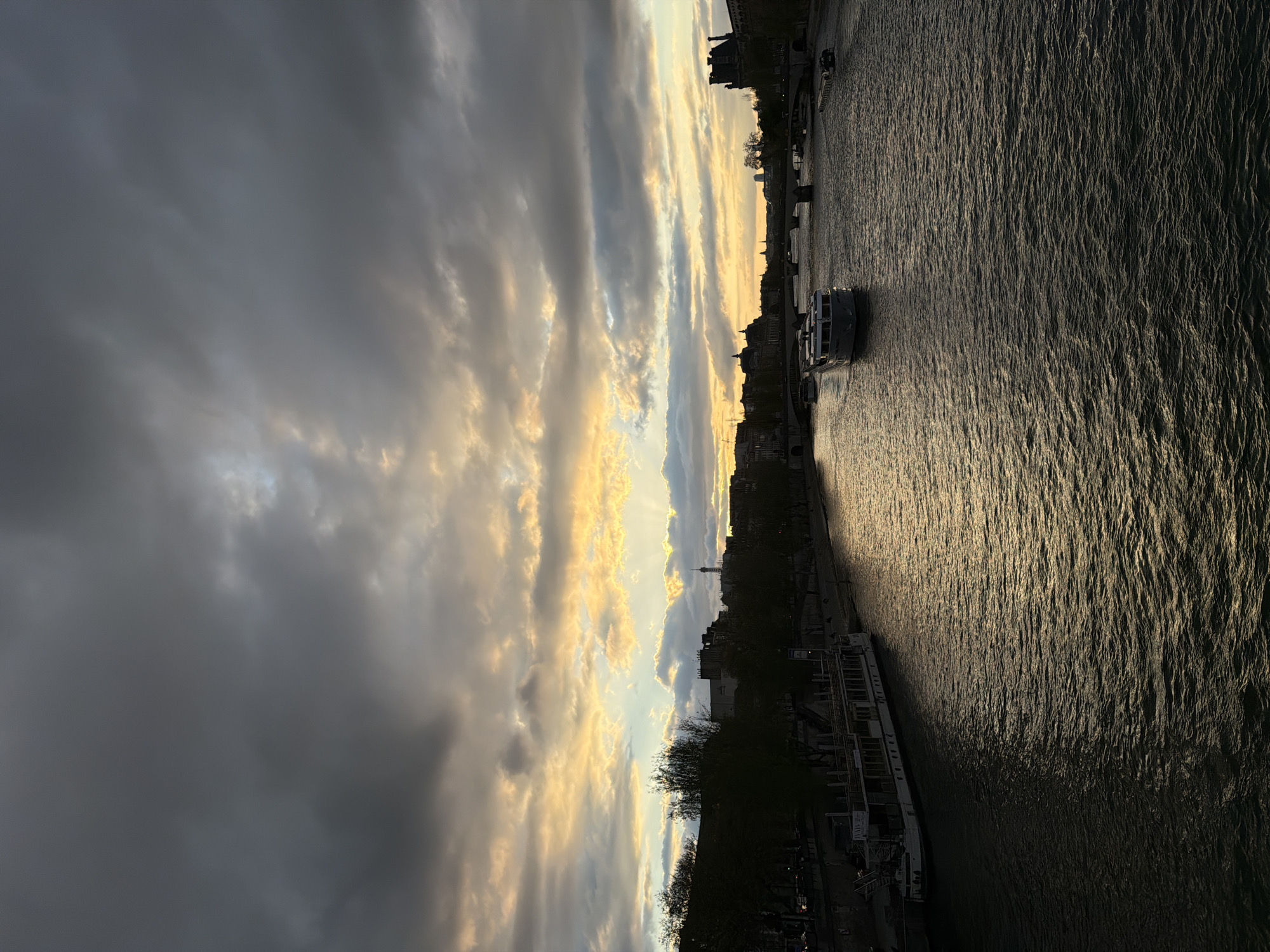 The image depicts a view of the Seine River in Paris, France, at dusk or dawn. The sky is filled with dramatic clouds, illuminated by a golden light from the sun. The Eiffel Tower is visible in the distance, partially obscured by buildings. A tour boat is present on the river, and docked boats line the riverbank. Trees and buildings are silhouetted against the sky.