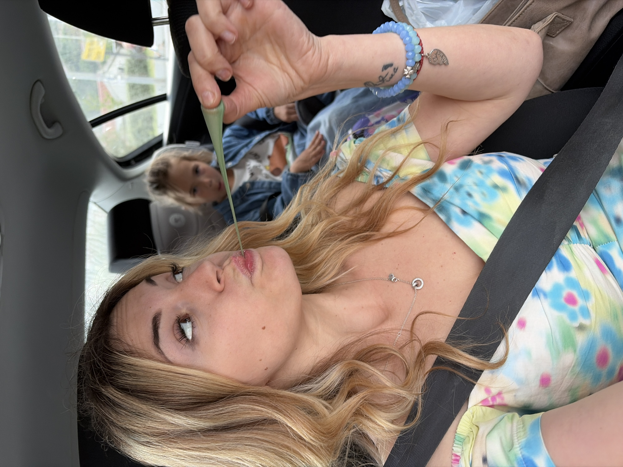 A playful, candid close-up shot captures a blonde woman inside a car, possibly in Budva, Montenegro. She is the central focus, positioned in the passenger seat with a black seatbelt diagonally across her chest. She wears a vibrant tie-dye top with blues, greens, yellows, and pinks, and a silver necklace with two interlocking circle charms. On her right wrist, she wears a blue beaded bracelet adorned with a small silver charm and a red string detail. A small, indistinct tattoo is visible on her left forearm.
The woman is shown engaging in a lighthearted activity, holding a long, light green, gooey substance (likely slime) with her right hand, bringing it close to her slightly puckered lips in a humorous expression. Her long, wavy blonde hair cascades over her shoulders.
In the blurred background, a young blonde child, wearing a denim jacket over a white top, is visible, suggesting they are also in the car. The car interior features a gray headliner and a grab handle. Light streams in through the car windows and a sunroof, indicating it is daytime. A blurred outdoor scene, possibly of buildings or a city environment, is visible through the windows. A brown bag or purse is also partially visible in the background. The image conveys a sense of fun and spontaneity.