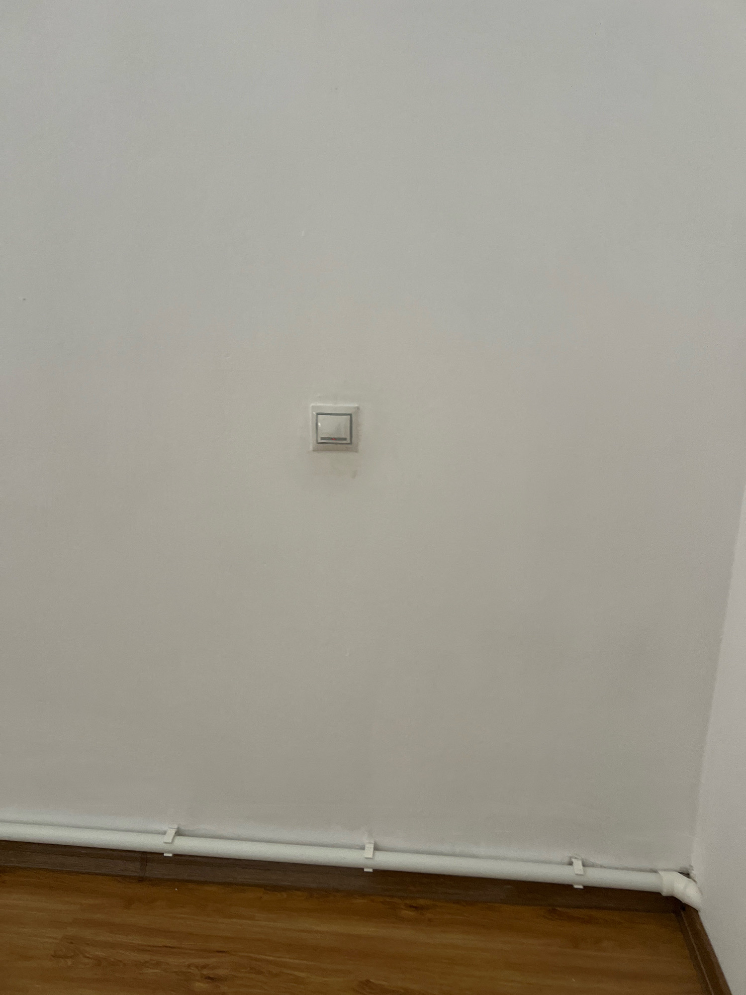 A light switch is mounted on a white wall. Below the switch, a white pipe runs horizontally along the base of the wall, attached by white clips. The floor is made of wooden planks. The scene is indoors, with no specific geographical indicators to place it in Novogagatli, Russia. There are no people or actions depicted.