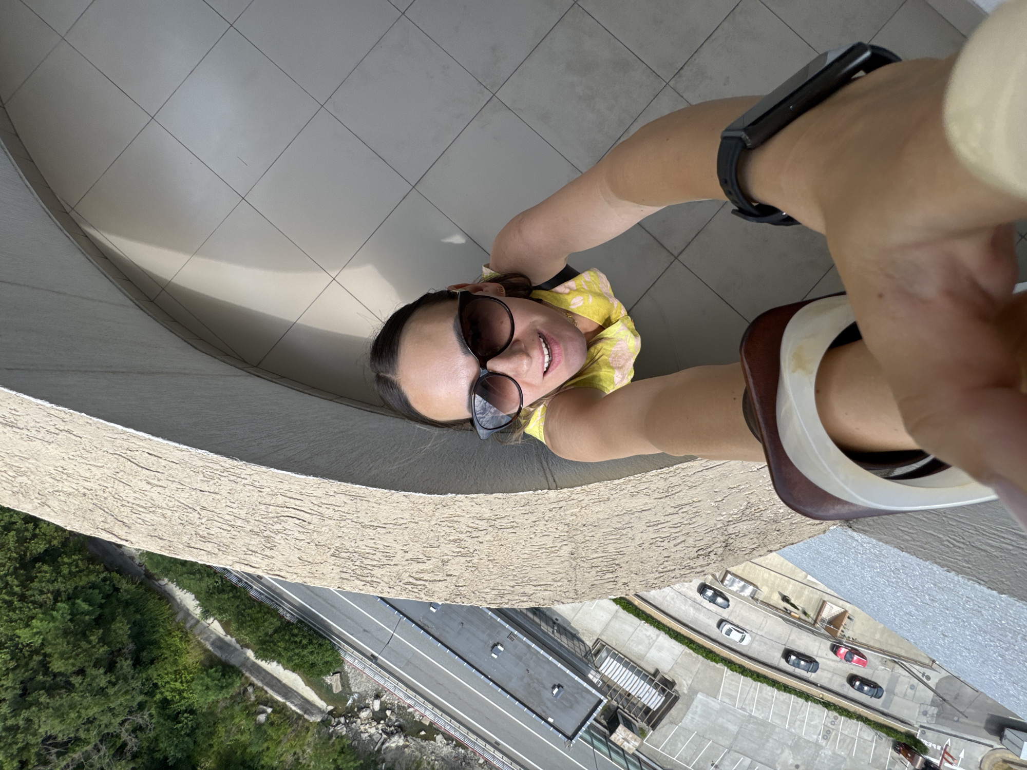 A first-person perspective selfie captures a young woman standing on a balcony, looking down and smiling. She is wearing sunglasses, a yellow floral top, and a black fitness tracker on her left wrist. Her legs are bent at the knees, and she is holding the camera with her right hand, which is partially visible in the frame. The background shows a tiled floor and a textured wall. Below the balcony, a road with cars can be seen, surrounded by lush green trees and some rocky terrain. The scene suggests a bright, sunny day, possibly in the late morning or early afternoon, given the strong shadows cast by the sunlight. The location is identified as Sochi, Russia. The woman appears happy and is enjoying the view from her elevated position. No visible text is present in the image.