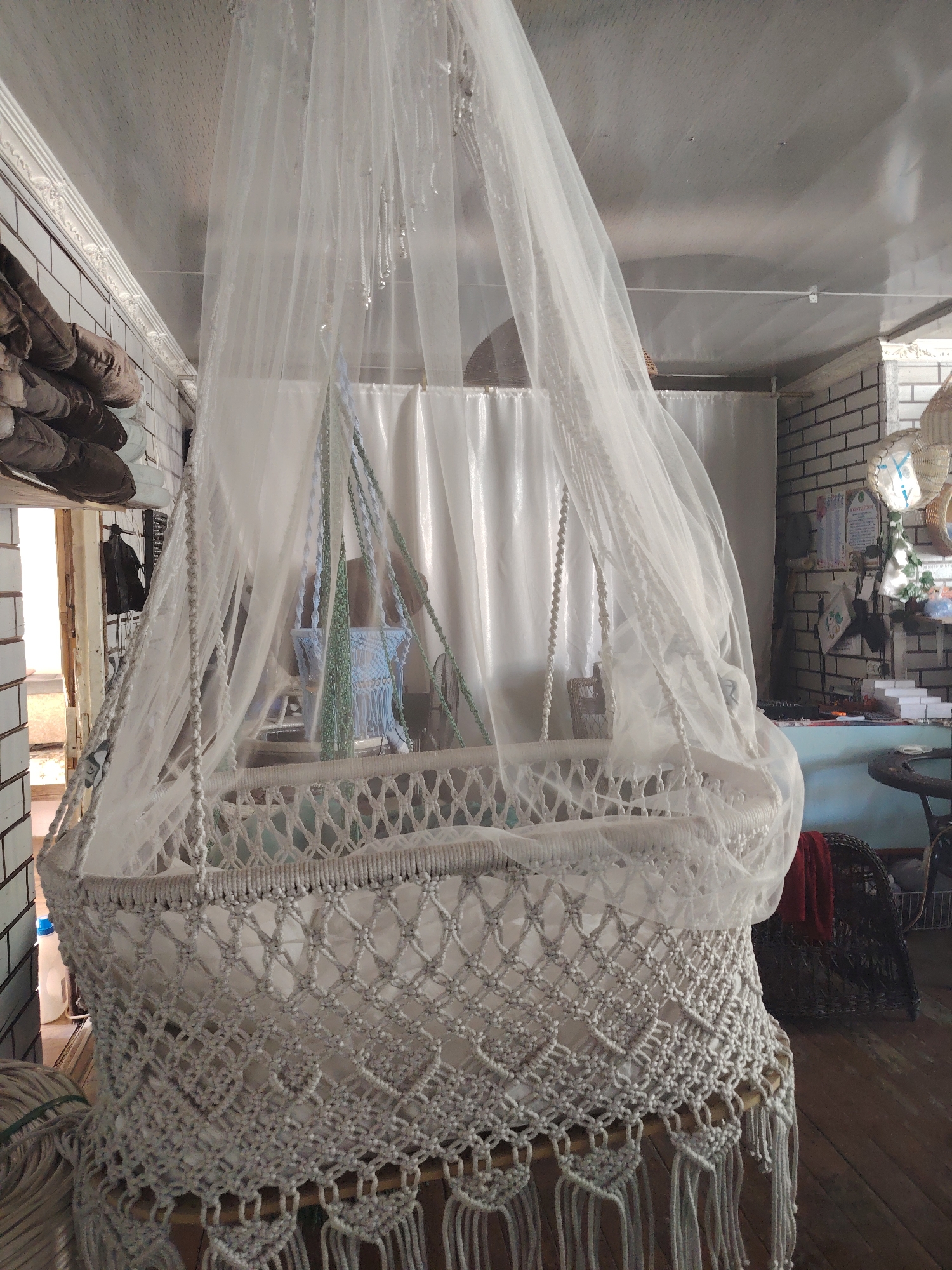 This image depicts a macrame baby bassinet hanging from the ceiling, covered by a sheer white canopy. The bassinet is intricately woven with white cords, featuring a lattice pattern and decorative fringes at the bottom. Suspended from above, green and blue cords are visible, likely part of the hanging mechanism. The background shows a room with white tiled walls on the left, a stack of what appear to be shoes on a shelf, and a large white curtain. On the right, a brick wall is visible, adorned with various items, including pictures and woven decorations. A dark woven chair with a red cloth draped over it is also present in the background, along with a round table. The floor is made of wooden planks. The overall scene suggests an indoor setting, possibly a home or workshop, in the city of Ibrat, Uzbekistan. No people are visible in the image.
