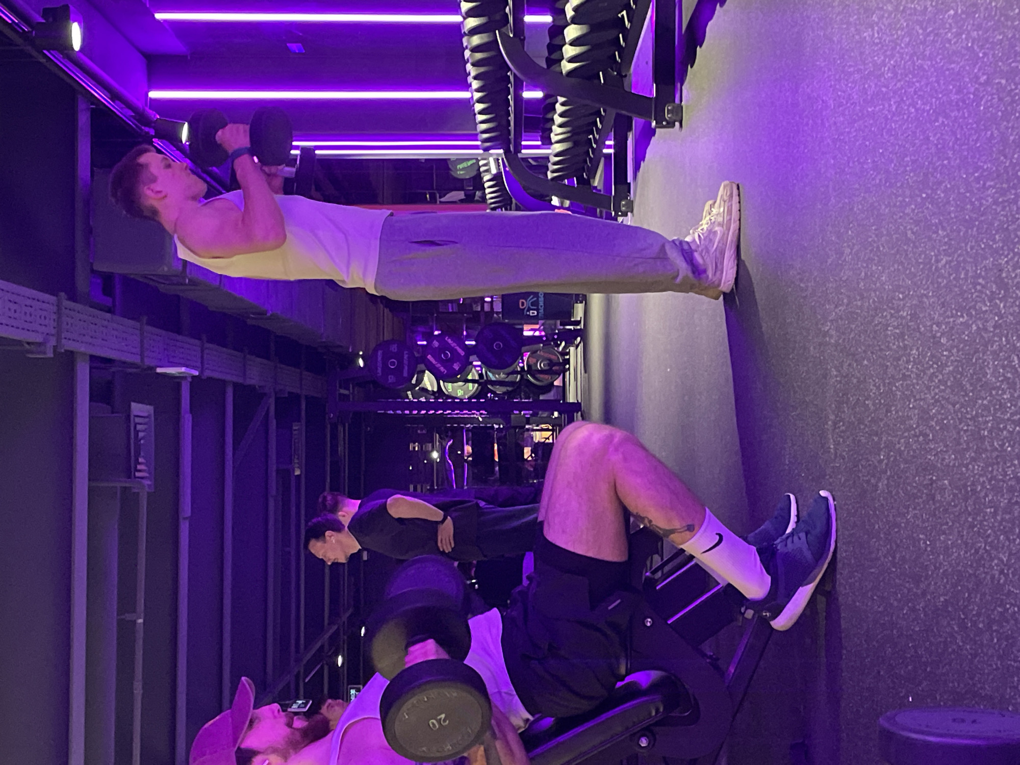 The image shows the interior of a gymnasium, illuminated by purple lighting. In the top part of the frame, a man in a white tank top and gray sweatpants is standing and lifting dumbbells. His feet are touching the wall behind him and he has a focused expression on his face. Below him, another man is seated on a workout bench, lifting a dumbbell labeled "20." The man is wearing a white tank top, black shorts, and a pink baseball cap. His legs are bent and positioned on the foot support of the bench. Another person in dark clothing is standing behind the man on the bench, seemingly observing or assisting. Racks of weights and workout equipment are visible in the background. The environment appears to be a modern and well-equipped fitness center. The city is not evident from the image.