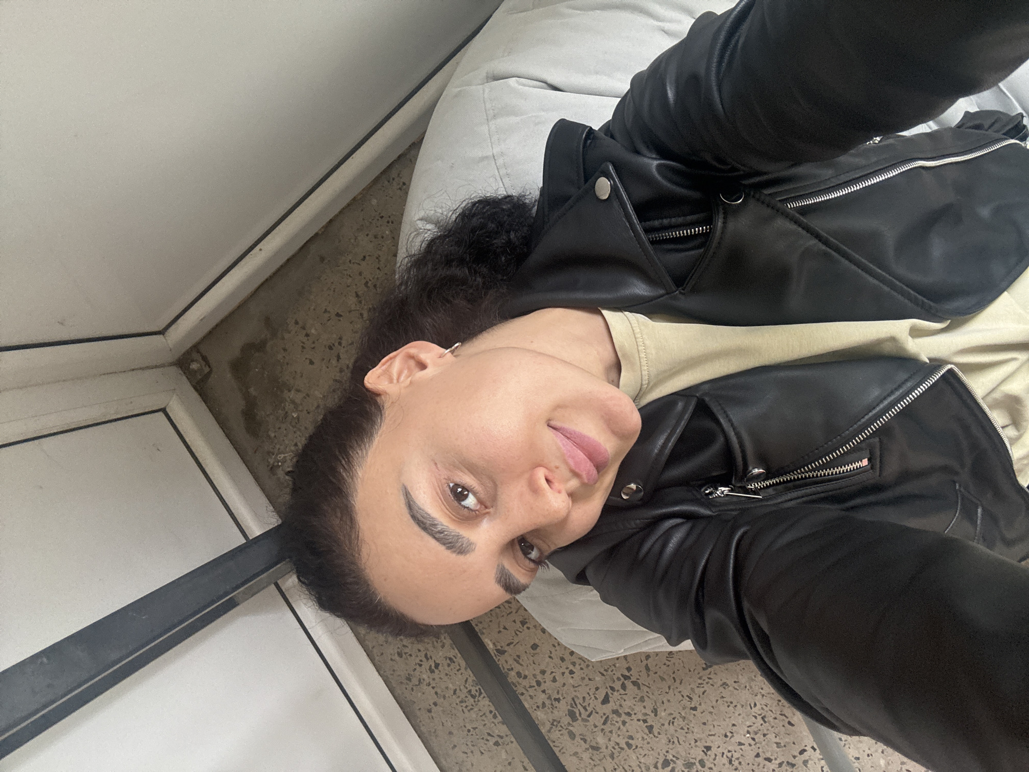A woman is lying on a white beanbag cushion, wearing a black leather jacket over a light-colored shirt. She is indoors next to a white wall with a black trim. The floor is a light beige with darker spots. The location appears to be in the city of Tbilisi, Georgia.