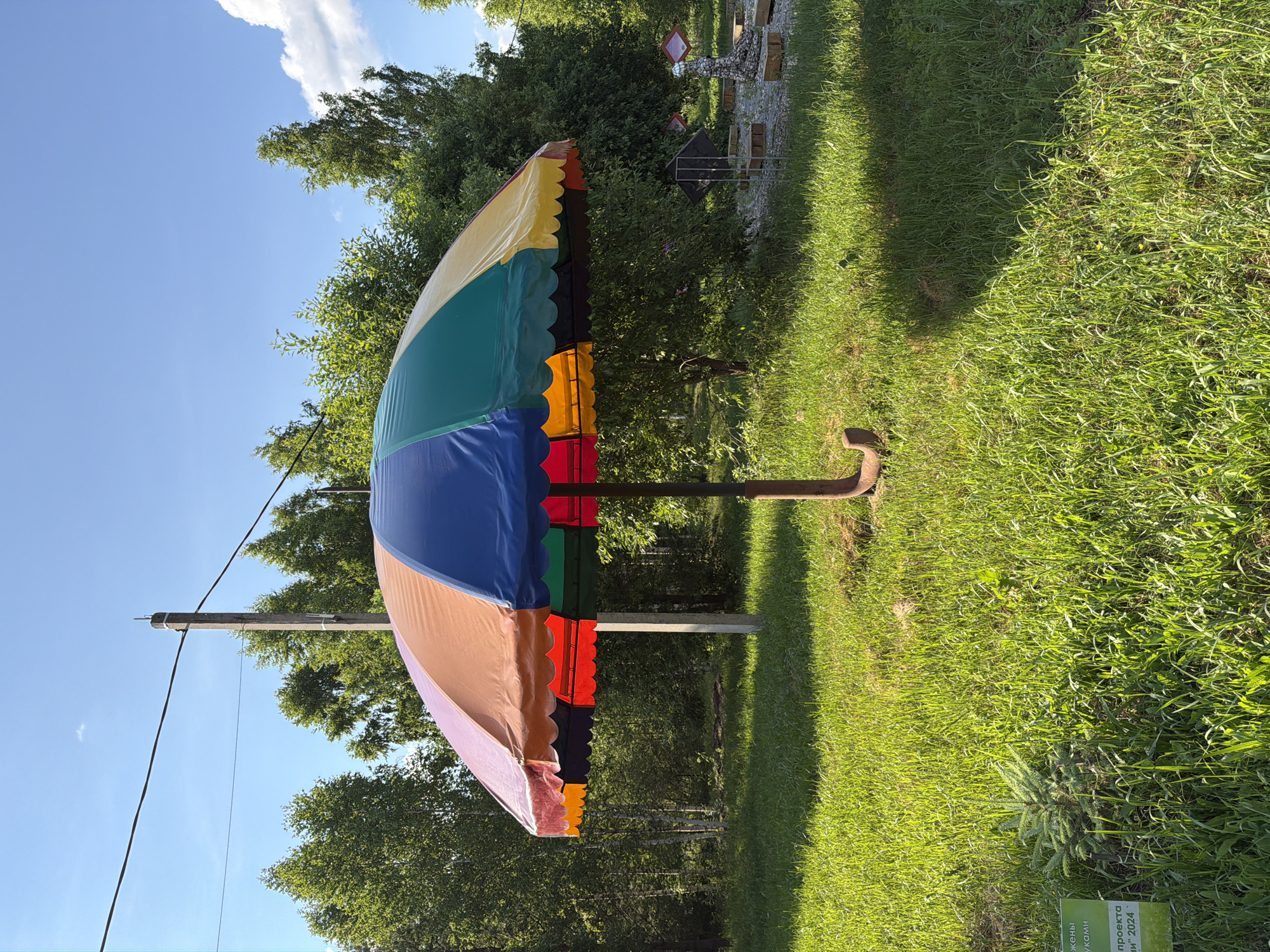 In the city of Zhirnosovo, Russia, a large, multi-colored umbrella stands planted in a grassy area. The umbrella's canopy is divided into segments of orange, red, dark blue, green, and yellow, with a scalloped edge. Its pole is made of a rusted metal pole, anchored by a curved base. Behind the umbrella, a concrete utility pole with wires stretches across the frame, and a dense forest of green trees forms the background. The sky above is a clear, bright blue with scattered white clouds. In the foreground, lush green grass covers the ground, with a small evergreen sapling visible to the left, next to a green sign with text in Russian. In the middle ground to the right, there are a few objects that appear to be sculptures or decorative elements, along with wooden blocks and a dark diamond-shaped object.
