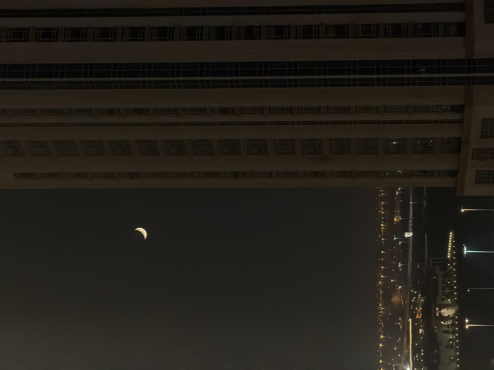 A crescent moon hangs in the dark night sky above an illuminated cityscape. To the right, the side of a tall, modern building with many windows dominates the frame. The building's facade is a light beige color, with vertical dark blue glass sections and horizontal rows of windows. Below the prominent building, the city stretches out, a sprawl of lights from buildings, street lamps, and possibly vehicles. Several distinct buildings with arched doorways and illuminated windows are visible in the foreground of the cityscape. The overall impression is one of urban nighttime activity under a celestial glow.