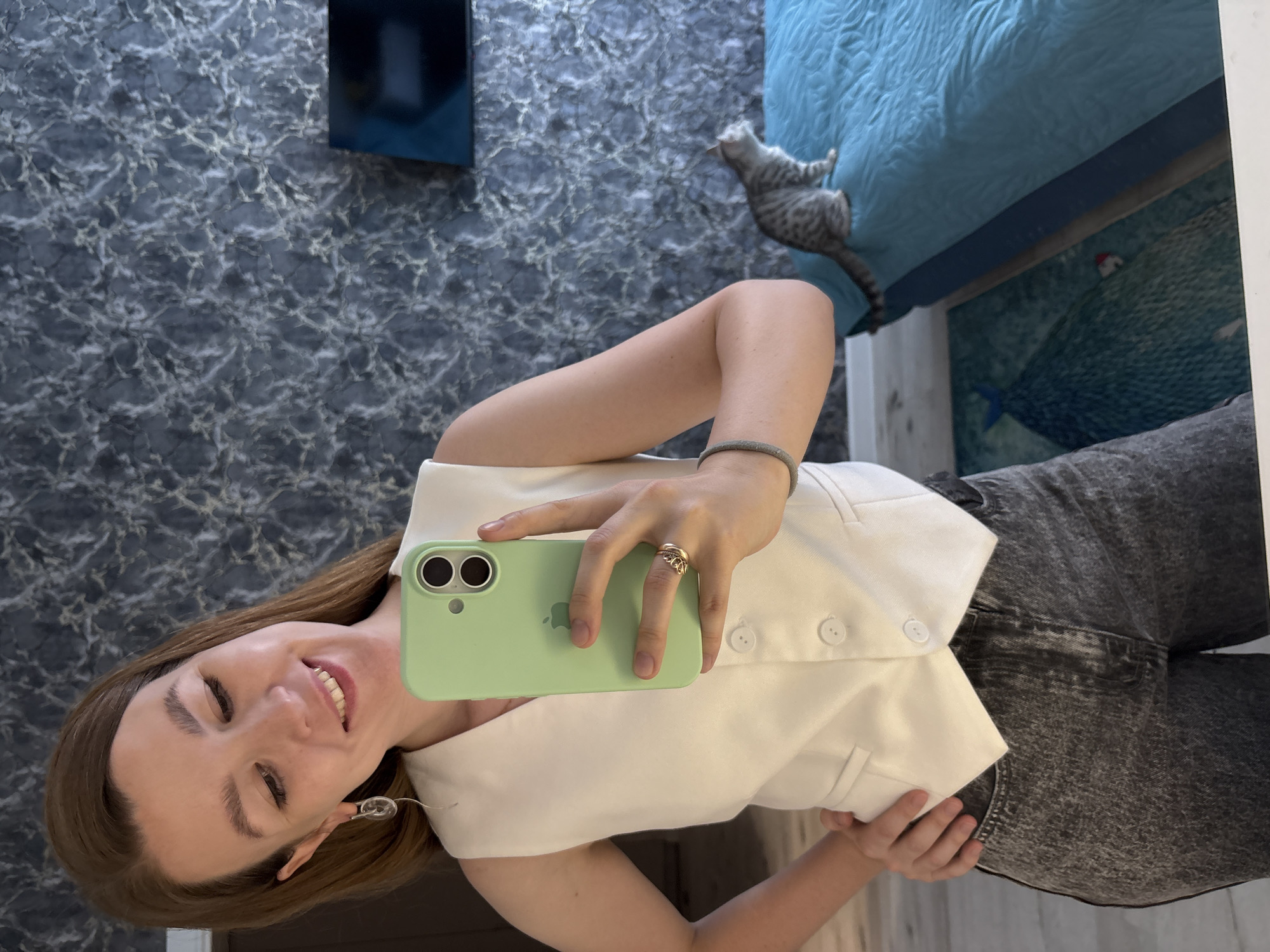 A woman with light brown hair is taking a selfie in a room with dark, patterned wallpaper. She is wearing a white sleeveless vest and dark, acid-washed jeans. She is holding a light green smartphone in her right hand, which is positioned to capture the photo. Her left hand is resting on her hip. A grey tabby cat is visible in the background, sitting on a bright blue quilted bed. A portion of a television screen is mounted on the wall. A rug with a blue and green design is on the floor. The image appears to have been taken indoors, likely in a residential setting.