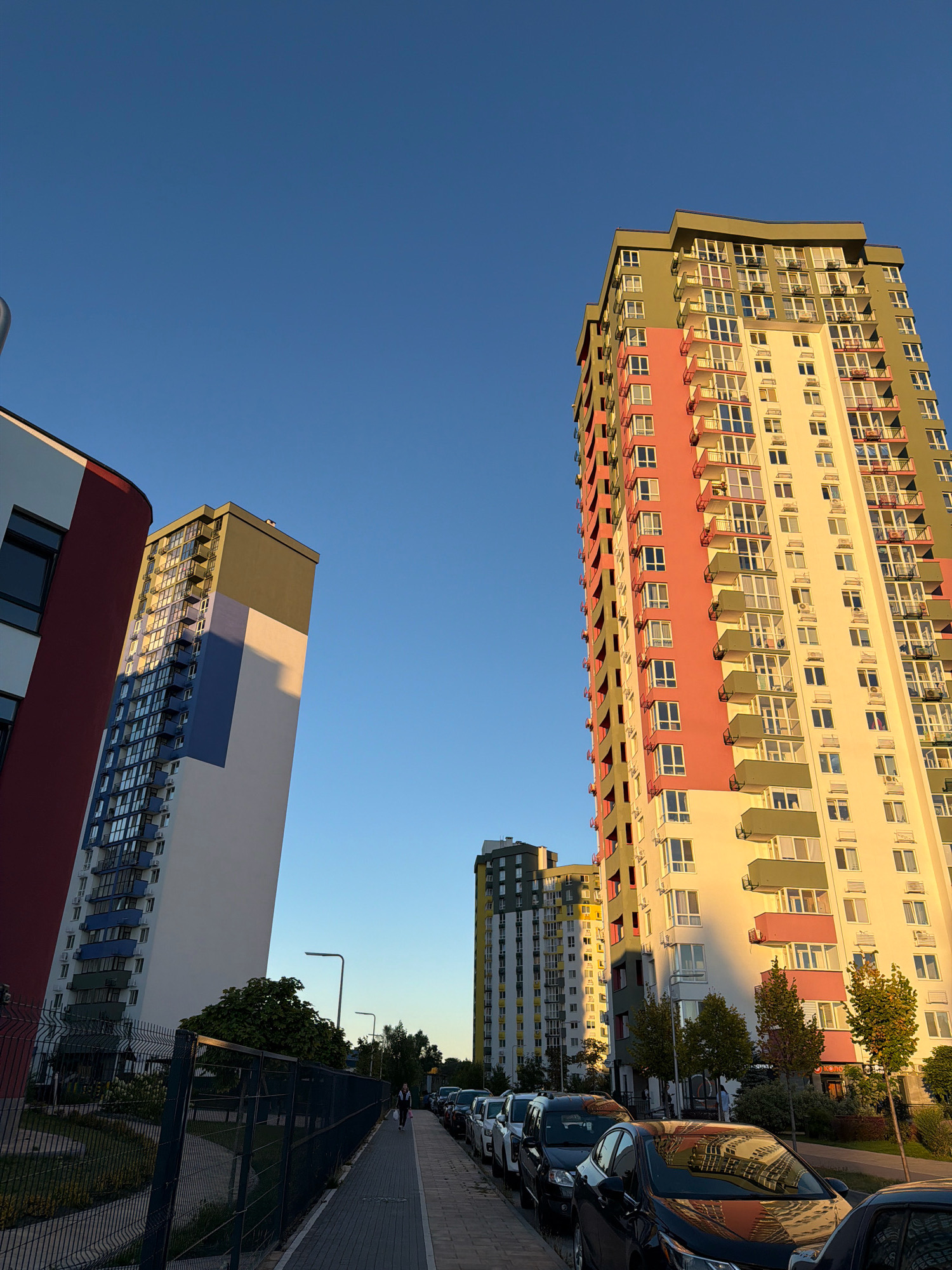 The image depicts a street scene in Kyiv, Ukraine, featuring a row of modern, multi-story apartment buildings. The buildings are characterized by their colorful facades, with sections painted in shades of cream, pink, green, and blue. Balconies with railings are visible on many floors. The buildings are tall and appear to be part of a residential complex.
In the foreground, a sidewalk runs alongside the buildings, lined with parked cars. The cars are of various makes and models, mostly dark in color. A black car in the immediate foreground reflects the colorful buildings and the blue sky.
A dark-colored metal fence runs along the edge of the sidewalk, separating it from a landscaped area with trees and grass. A person is walking away from the camera on the sidewalk, dressed in dark clothing and carrying a backpack. Streetlights are also visible along the sidewalk.
The sky is a clear, deep blue, indicating a sunny day. The low angle of the sun suggests it is either early morning or late afternoon, casting long shadows on the buildings and the ground. The overall impression is one of a contemporary urban residential area in Kyiv.