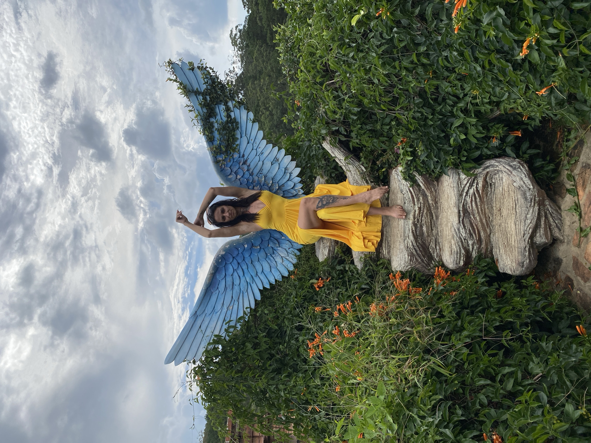 A woman in a yellow dress sits on large, textured rocks. She has her arms raised above her head, with her hands behind her neck, and her legs are crossed. Behind her are large, blue, feathered wings, which appear to be part of a sculpture. The wings are adorned with green vines. The woman is surrounded by lush green foliage and orange flowers. In the background, there are trees and a cloudy sky. This scene is located in Cam Ly, Da Lat, Vietnam.