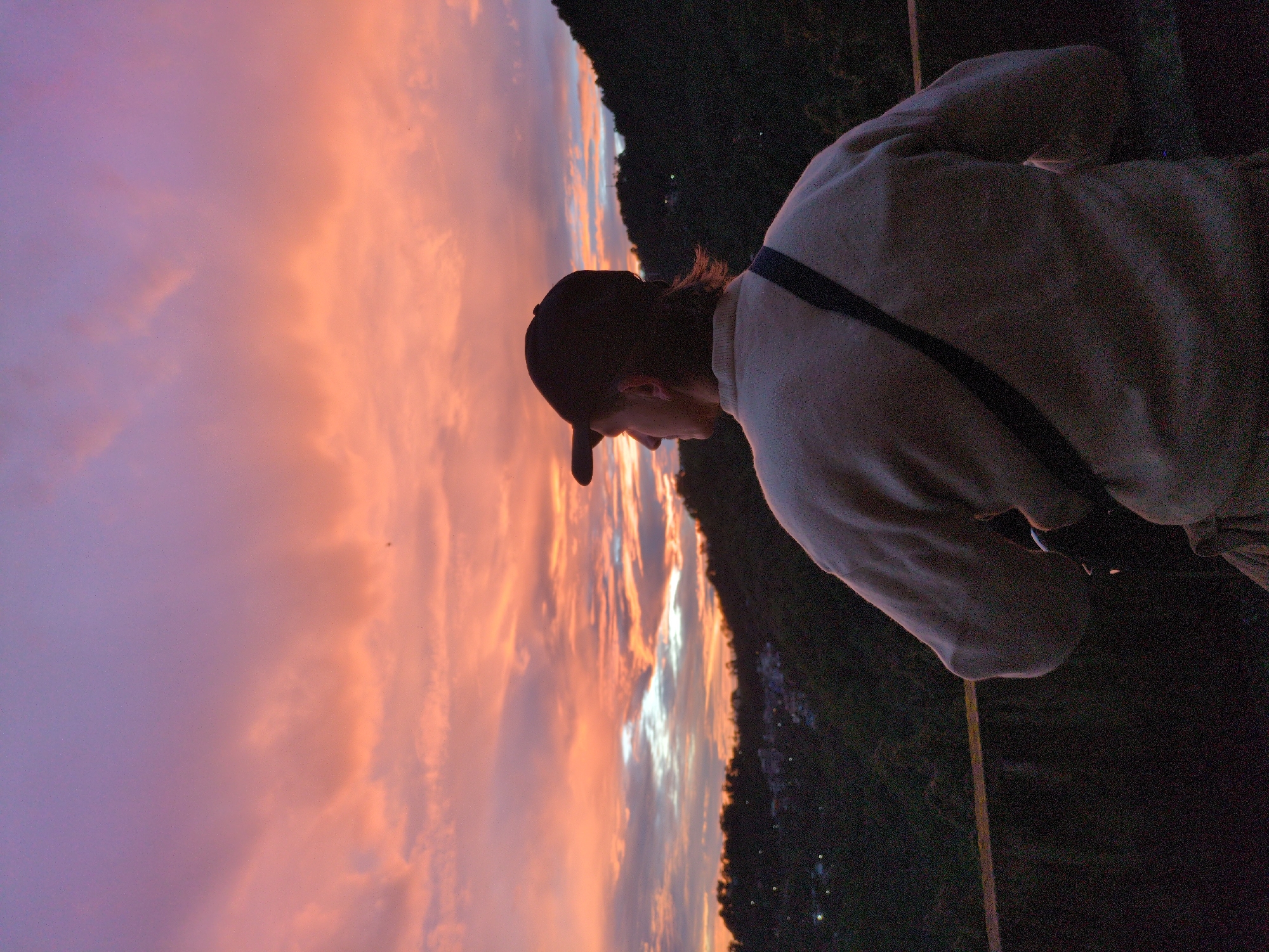 The image shows a person from behind, wearing a light-colored sweater and a baseball cap, looking out at a vibrant sunset over a hilly landscape. The sky is filled with dramatic clouds painted in hues of pink, orange, and purple, indicative of the twilight hours. In the background, the slopes are covered in dark green vegetation, with scattered lights suggesting a settlement in the valley below, characteristic of Dharamkot, India. The person is standing on what appears to be a balcony or viewing area, with a railing in the foreground. The overall mood is one of peaceful contemplation of the natural beauty.