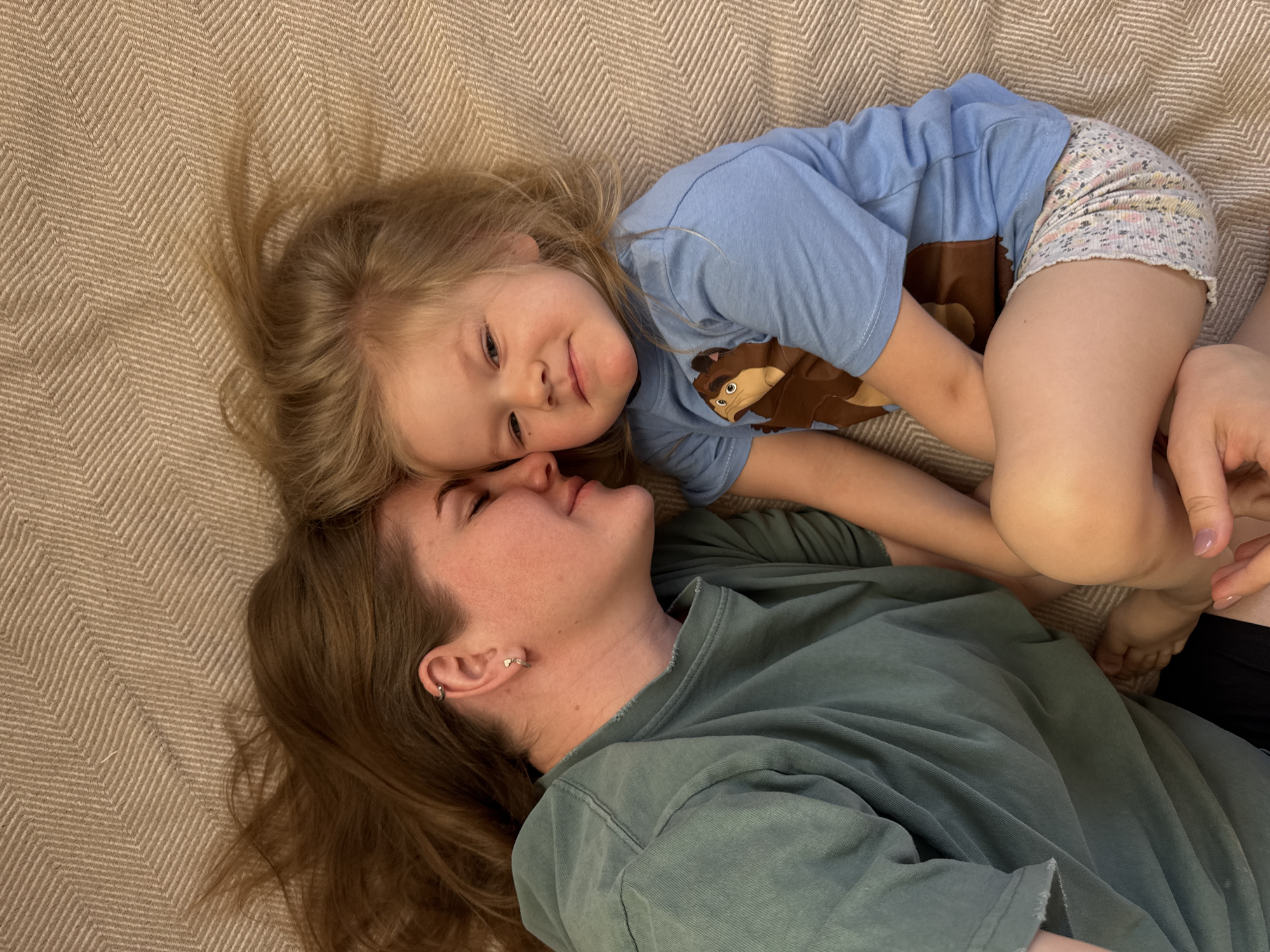 A woman and child are lying on a tan herringbone patterned blanket. The woman is lying on her back facing upward with her head to the left side of the frame, with her light brown hair spread around her head. She is wearing a green t-shirt and has a small silver hoop earring in her left ear. A young child is lying on top of her, turned facing her. The child has blond hair spread around her head. She is wearing a light blue t-shirt with a cartoon animal on it and floral patterned shorts. Her right arm is over the woman's arm. Both are looking at each other. The location is unknown, but it is stated to be in the city of Ферма, Russia.