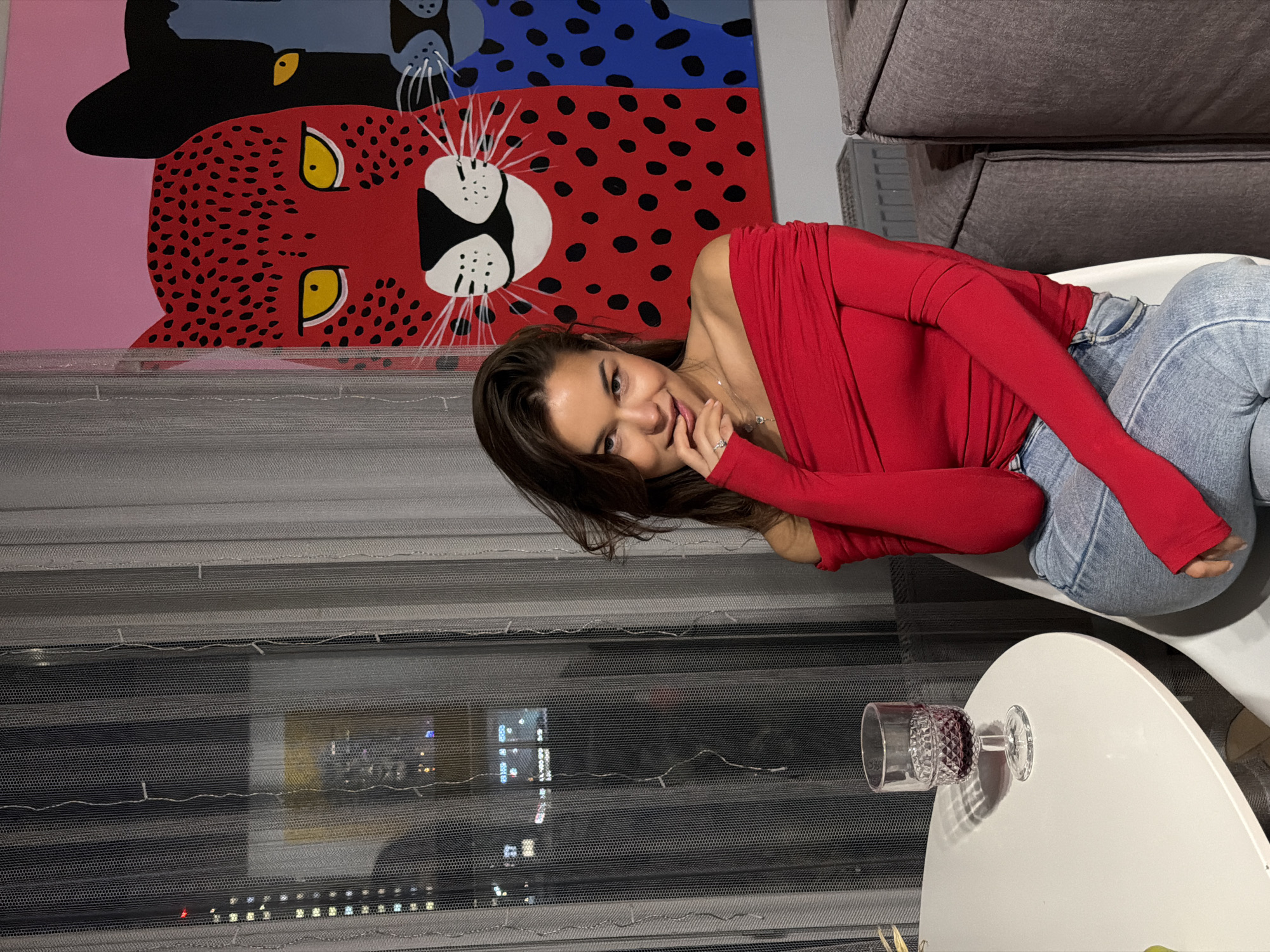 A young woman with long, dark hair reclines on a light-colored chair or sofa in an indoor setting, likely an apartment in Krasnoyarsk, Russia. She wears a vibrant red, long-sleeved, off-the-shoulder top and light blue jeans. Her expression is a soft, inviting smile, and she looks directly at the viewer, with one hand gently touching her chin or mouth.
Behind her is a large, colorful painting featuring two stylized animal faces. The prominent lower face is a red, leopard-like creature with black spots and bright yellow eyes on a pink background, while an upper face is a blue creature with black spots and yellow eyes on a blue background. Both have distinct white snouts and whiskers. To the right, a dark grey upholstered sofa is partially visible.
In the foreground, a round white table holds a single cut glass goblet containing a dark liquid, possibly wine. The scene is set at night, indicated by the dark outside view through horizontal window blinds or sheer curtains. Blurred lights of an urban cityscape are visible through the window, suggesting a high-rise view. The overall atmosphere appears relaxed and comfortable. There is no visible text in the image.