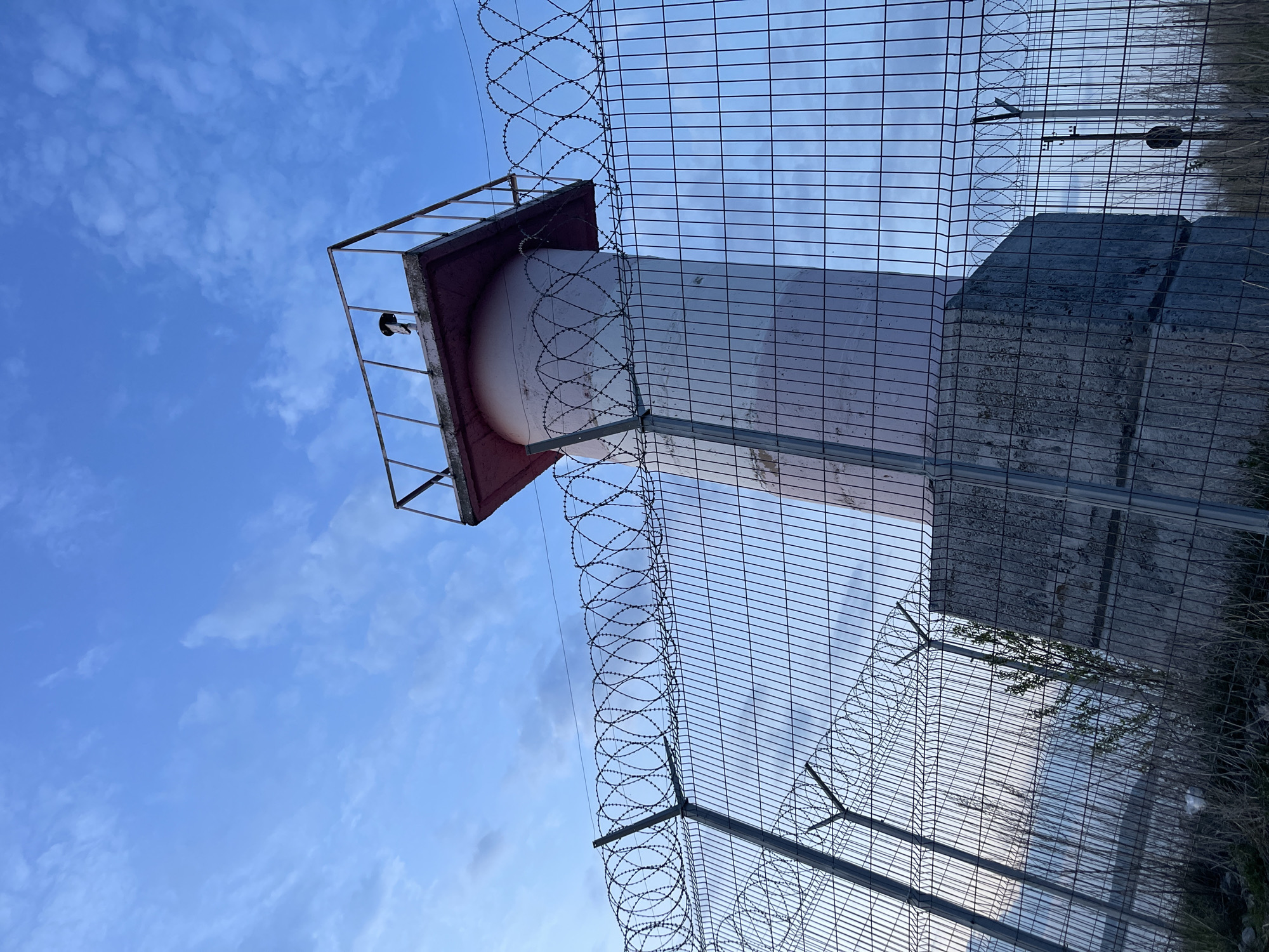 The image depicts a water tower located in what is believed to be the city of Chaikovsky, Russia. The tower is cylindrical with a spherical top, painted primarily white with a maroon-colored top. It's surrounded by a metal security fence topped with barbed wire. The sky is light blue with scattered clouds. The tower seems to be within a fenced-off area.