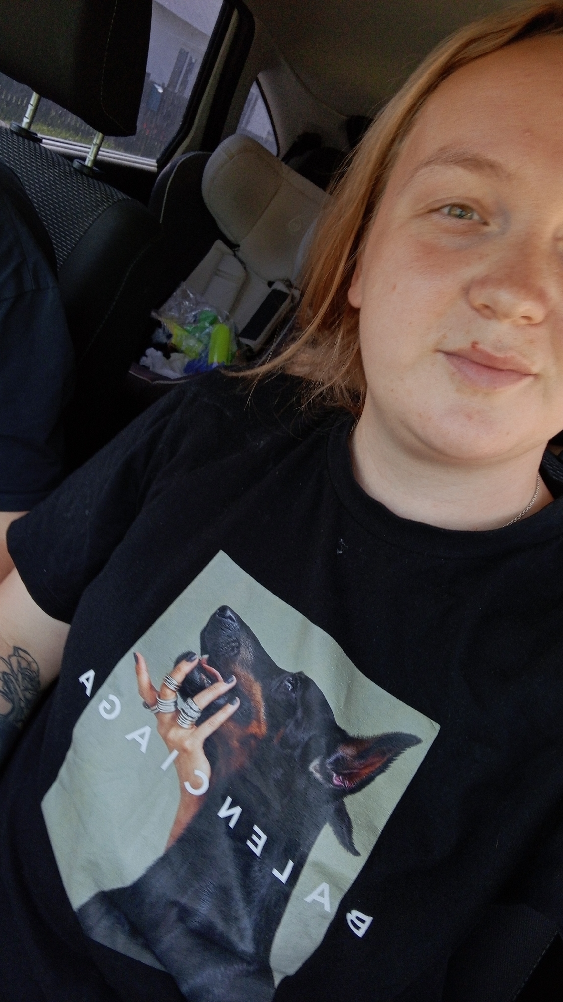 A woman is seated in a car, presumably in the city of Уральское, Russia, based on the prompt instructions. She is wearing a black t-shirt with a print of a Doberman dog and a hand adorned with rings. The interior of the car includes black seats, a child safety seat, and various items in the back. The woman is facing the camera, suggesting she may be taking a selfie.