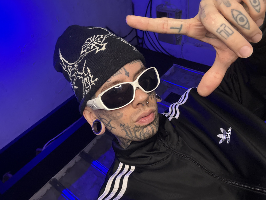 A heavily tattooed individual, appearing male, is depicted in a medium close-up, posing against a dark background illuminated by blue light. The subject wears white-framed sunglasses with dark lenses, a black knitted beanie featuring white abstract designs, and a black Adidas tracksuit jacket with three white stripes on the sleeves and the trefoil logo on the left chest. Prominent facial modifications include large ear gauges (tunnels) in both earlobes, a nasal septum ring, and two nostril studs. Extensive black ink tattoos cover the individual's face, neck, and visible hands. The individual's right hand is raised, forming an "L" shape with the index finger extended vertically and the thumb extended horizontally. Visible tattoos on the fingers of the right hand include a "T" on the index finger, an "F" and "2" on the middle finger, and an "eye" symbol on both the ring and pinky fingers. The blurred background suggests an indoor or semi-enclosed industrial-style space with structural beams and electrical conduits visible, all cast in a dominant blue hue. The specific location within the city of Buenos Aires, Argentina, cannot be determined from the visual evidence provided.