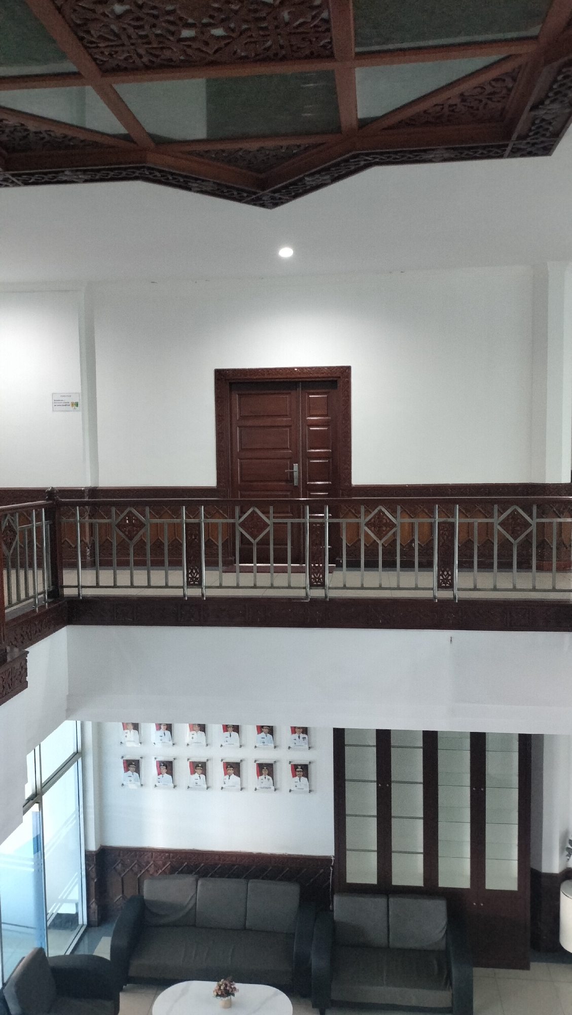 This image depicts the interior of a building, likely a government office or educational institution, in Lhokseumawe, Indonesia. The perspective is from a higher floor looking down into a central atrium.
In the foreground, on the lower level, are two dark-colored sofas arranged around a white coffee table with a small floral centerpiece. A portion of another sofa is visible on the right. To the left, a large window or glass partition allows a view of the outside. The wall behind the seating area features a decorative wooden paneling and a row of framed portraits of men, arranged in two rows of six. To the right of the portraits is a tall, dark wooden cabinet with multiple glass doors.
In the midground, a second-floor walkway is visible, featuring a dark wooden railing with intricate carvings and a metallic railing with a diamond pattern. A large, dark wooden double door is centered on the wall behind the walkway.
The ceiling is a prominent feature, with exposed wooden beams forming a grid pattern, some of which have ornate carvings. The walls are painted white. The overall impression is one of a well-maintained, somewhat formal interior space. The portraits suggest a connection to local leadership or important figures within the institution.