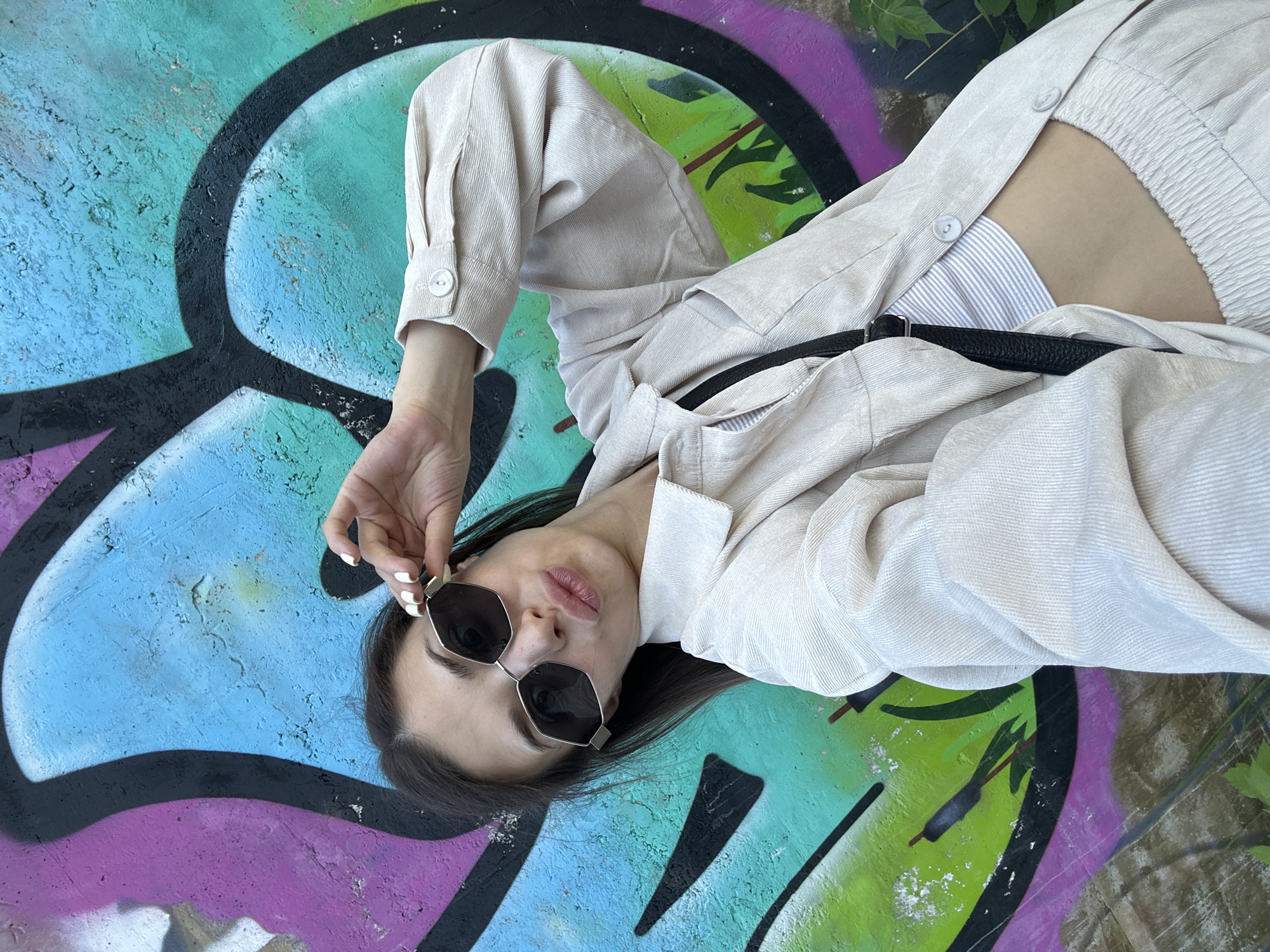 A young woman is lying on a colorful, graffiti-covered surface, possibly concrete, in Izhevsk, Russia. She is wearing a light-colored, corduroy-like jacket, a striped shirt underneath, and dark sunglasses. Her hand is raised, holding the frame of her glasses. A thin, black belt is visible. The graffiti consists of shades of blue, green, pink, and black paint.