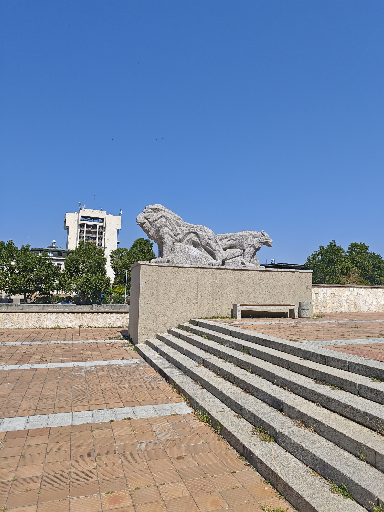 The image depicts the Lion's Bridge monument in Lovech, Bulgaria, under a clear blue sky. The monument features two stylized stone lions, one in front of the other, on a large, rectangular stone pedestal. To the right of the pedestal, a set of wide stone stairs leads down to a plaza paved with reddish-brown tiles and lighter grey accents. A stone bench and a trash receptacle are visible on the plaza level, in front of the pedestal. In the background, a modern multi-story building with a white facade and antennas on its roof stands amongst lush green trees. The overall scene is an outdoor public space, likely a square or park, on a sunny day.
