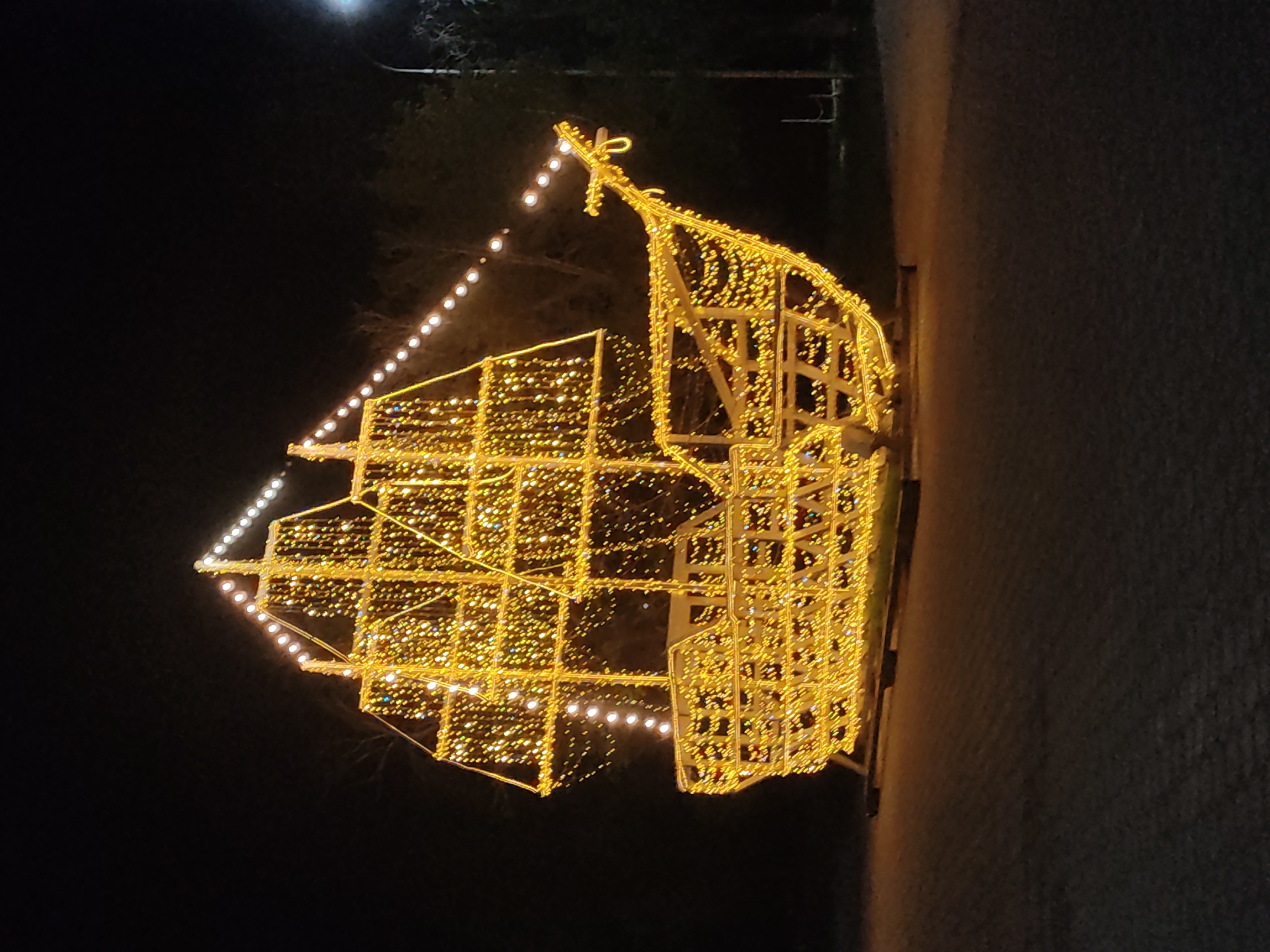 The image depicts a brightly illuminated structure in the shape of a ship at night. It's constructed with a frame covered in numerous small, warm-colored lights. The vessel is positioned on what appears to be a paved surface, next to a dark background. The location is Unknown, Unknown.