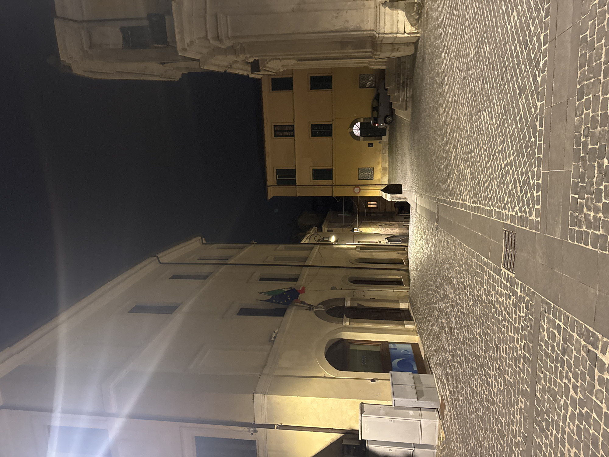 The image depicts a narrow street scene at night in Castelnuovo di Porto, Italy. The street is paved with cobblestones in a patterned design. Buildings flank both sides of the street, illuminated by dim streetlights. On the left, a multi-story building displays a flag, possibly the flag of Italy. The building's architecture features arched windows and a light-colored facade. The building on the right has square windows. A car is parked in the street. The sky above is dark. The image suggests an alley in an old section of town.
