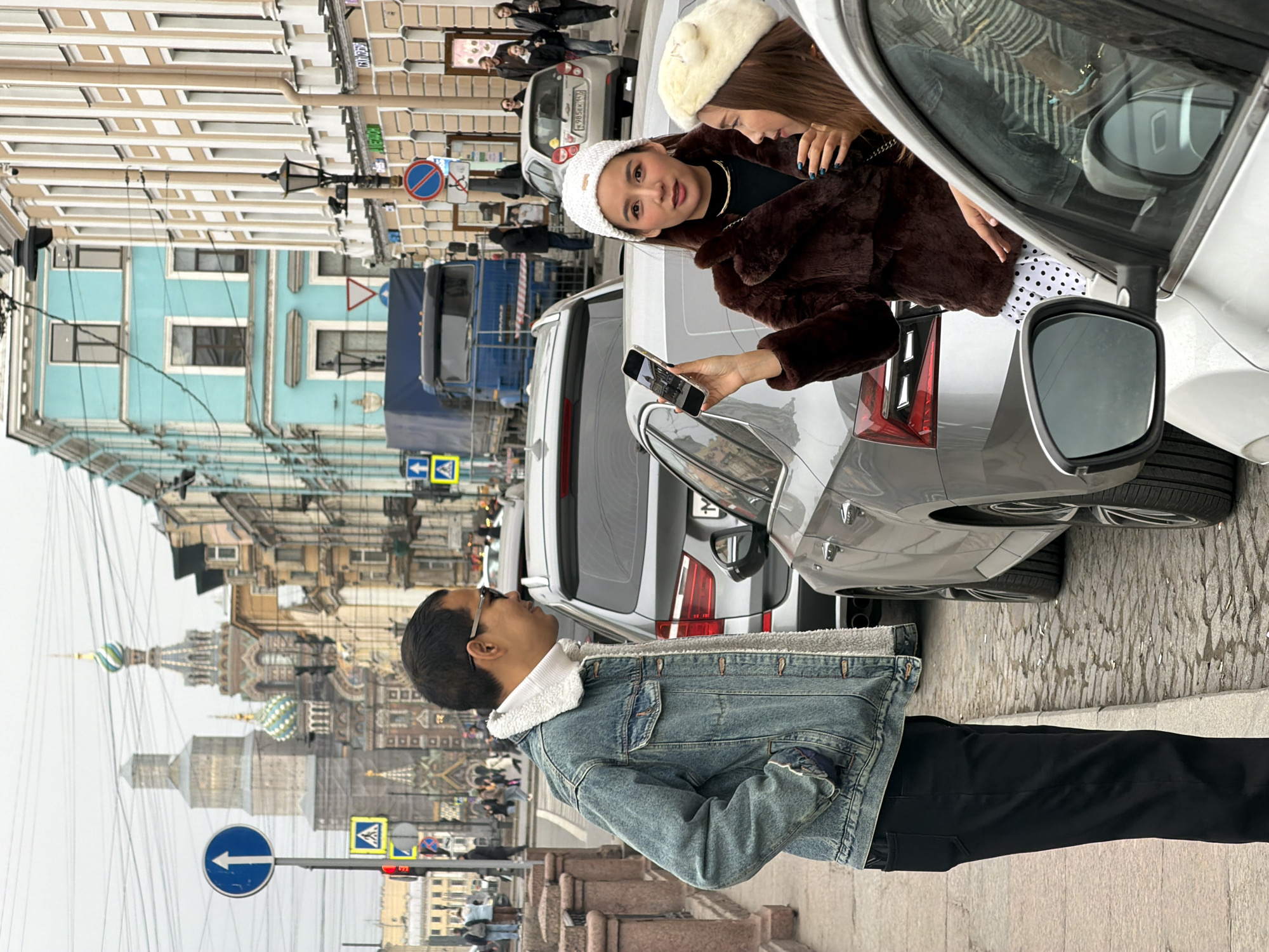 The image depicts a street scene in Saint Petersburg, Russia, on an overcast day, with the Church of the Savior on Spilled Blood prominently visible in the distant background, indicating the location near the Griboyedov Canal Embankment.
In the foreground, three individuals interact near parked vehicles. On the left, a male with dark hair, wearing sunglasses, a white turtleneck, a denim jacket with a white fleece lining, and black pants, stands on a concrete sidewalk with his right hand in his pocket, looking towards the right.
In the center-right, a female with long dark hair, dressed in a white knit beanie, a dark brown faux fur coat, a black top, and a white polka-dot skirt, holds a gold-colored smartphone in her left hand, displaying its screen. Her fingernails are painted blue, and she faces forward with her mouth slightly open. To her right, partially obscured by the side mirror of a white foreground vehicle, a second female with light brown hair and a cream-colored beret observes the smartphone screen.
Several cars are parked on the cobblestone street that runs parallel to the sidewalk. A silver sedan is positioned between the man and the women. Behind it, a dark grey SUV is visible. Further down the street on the right, a white sedan with Russian license plate "M 985 EK 178" and a blue truck are parked.
Architectural elements include multi-story historical buildings lining the street, featuring light brown, light blue, and beige facades. Street signage includes a blue circular "Ahead Only" arrow sign and a yellow triangular "Pedestrian Crossing" sign on a pole on the left. Further down on the right, a red circular "No Stopping" sign is visible. Overhead electrical wires crisscross the frame. The sky is uniformly overcast.