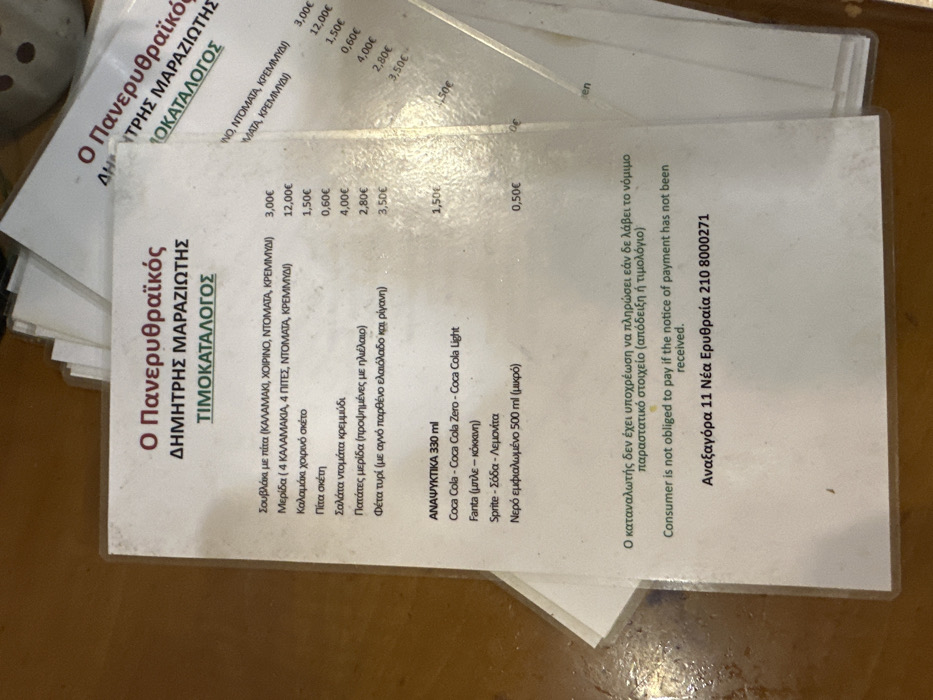 The image displays a physical menu, titled "ΤΙΜΟΚΑΤΑΛΟΓΟΣ" (Pricelist), from an establishment named "Ο Πανερυθραϊκός ΔΗΜΗΤΡΗΣ ΜΑΡΑΖΙΩΤΗΣ". The menu is primarily in Greek, with some English text for a legal disclaimer.
The menu lists various food items and their prices, including "Σουβλάκι με πίτα" (Souvlaki with pita) for 3,00€, a "Μερίδα" (portion) with skewers for 12,00€, "Καλαμάκι χοιρινό σκέτο" (plain pork skewer) for 1,50€, "Πίτα σκέτη" (plain pita) for 0,60€, "Σαλάτα ντομάτα κρεμμύδι" (tomato onion salad) for 4,00€, "Πατάτες μερίδα" (fries portion) for 2,80€, and "Φέτα τυρί" (feta cheese) for 3,50€.
A separate section for "ΑΝΑΨΥΚΤΙΚΑ 330 ml" (Soft Drinks 330 ml) includes Coca Cola, Fanta, Sprite, Soda, and Lemonade. "Νερό εμφιαλωμένο 500 ml" (bottled water 500 ml) is priced at 0,50€.
At the bottom, a disclaimer states in both Greek and English: "Consumer is not obliged to pay if the notice of payment has not been received." The contact information provided is "Αναξαγόρα 11 Νέα Ερυθραία 210 8000271", indicating the location in Nea Erythraia, Greece. The menu appears to be resting on a light brown surface, likely a table.