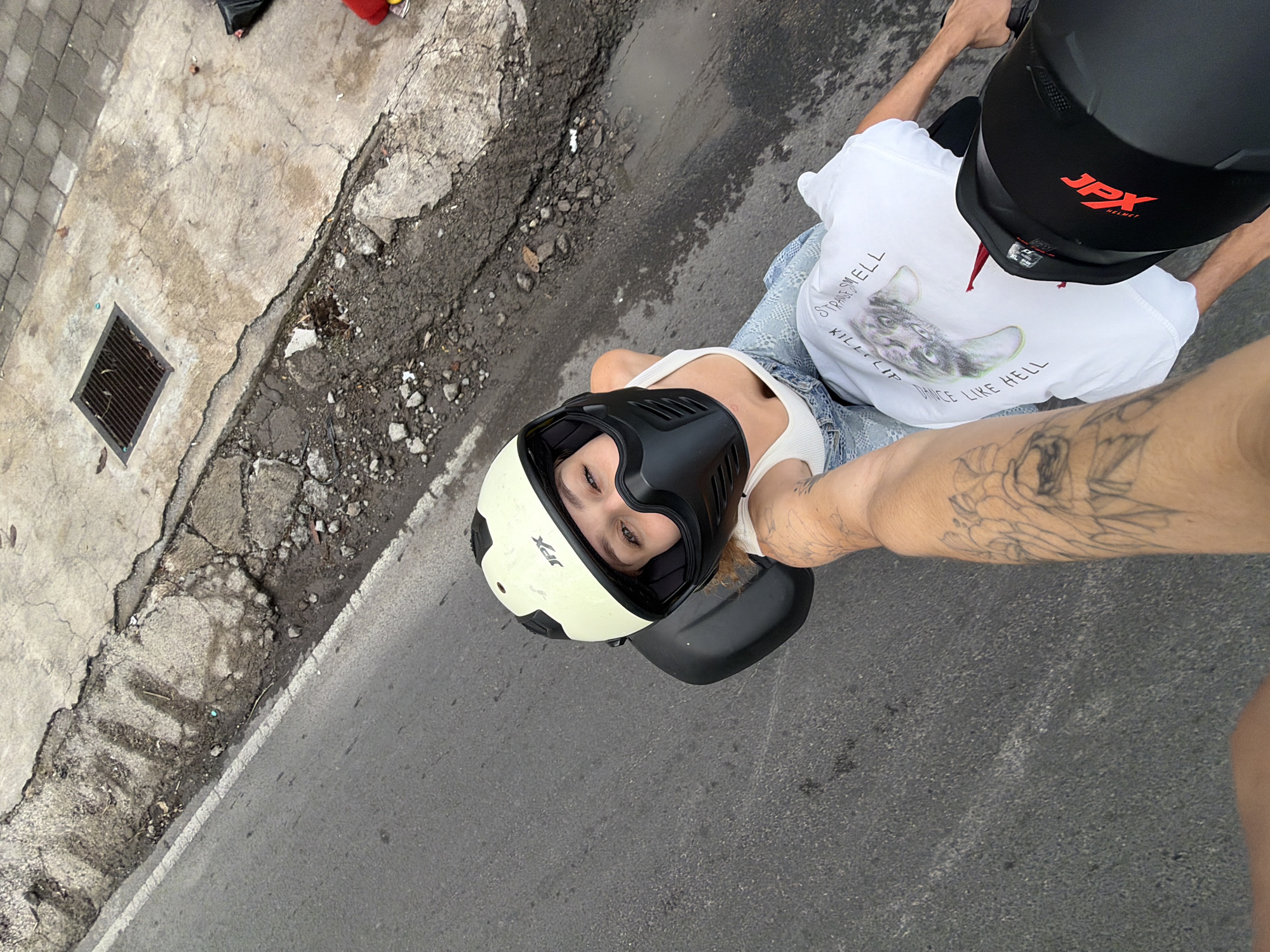 A first-person perspective selfie of a woman on a motorcycle in North Kuta, Indonesia. The woman, wearing a white tank top, denim shorts, and a light-colored helmet with a black face mask, is looking at the camera and smiling. Her left arm, adorned with a tattoo, is extended to take the photo. She is seated on a black motorcycle, with another black helmet visible in the foreground. The road beneath them is asphalt, with a cracked concrete sidewalk and a metal grate to the right. A white t-shirt with a graphic print and text is partially visible on her lap.