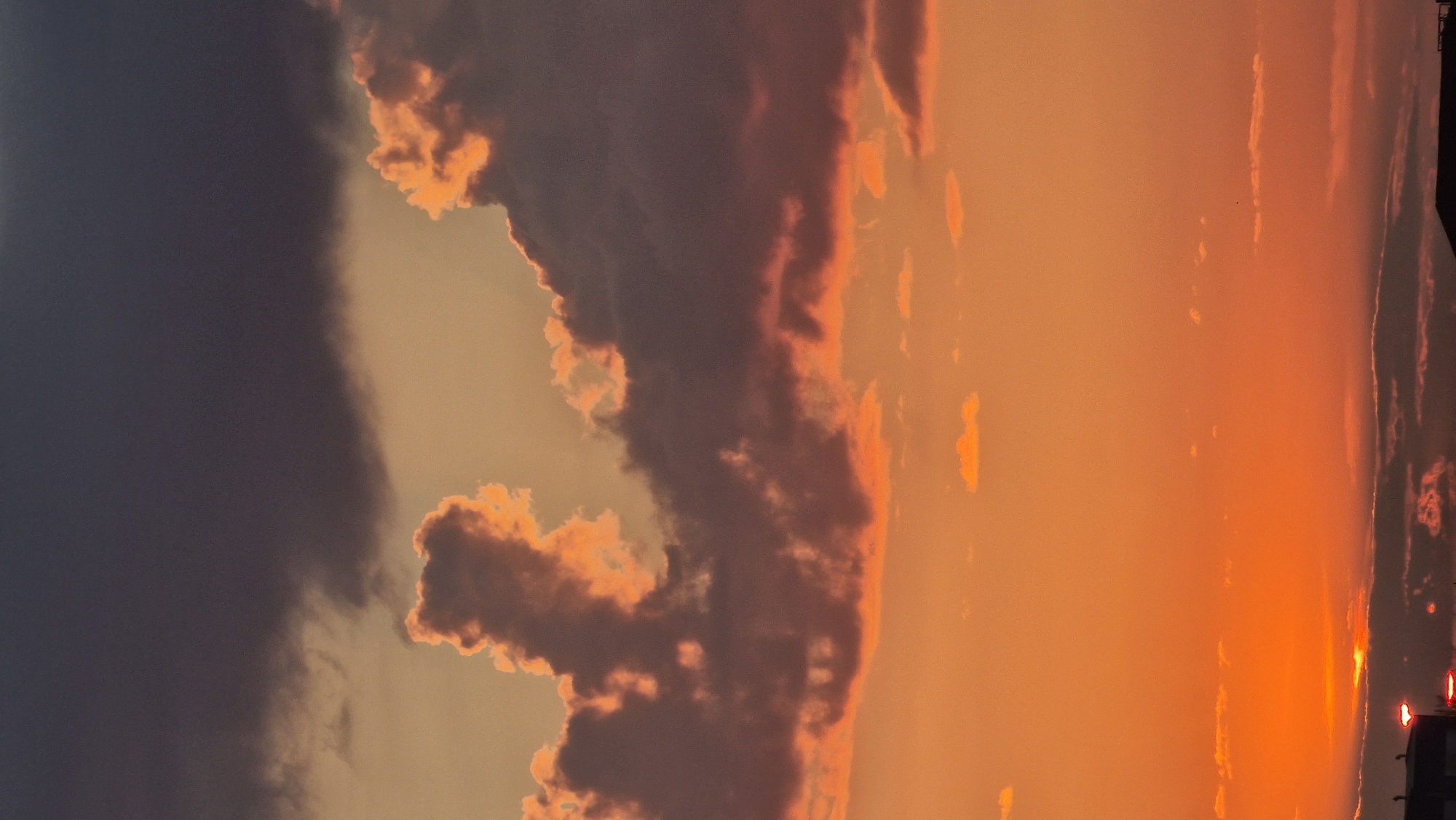 The image depicts a dramatic sunset over the city of Kostanay, Kazakhstan. The sky is a vibrant canvas of oranges, pinks, and purples, with dark, brooding clouds occupying the upper portion of the frame. These clouds are illuminated from below by the setting sun, creating a striking contrast. Smaller, scattered clouds are visible in the lower half of the sky, catching the warm hues of the sunset. In the foreground, the silhouettes of buildings and what appear to be streetlights are visible, suggesting an urban environment. The overall impression is one of a beautiful and intense natural phenomenon occurring over a city.