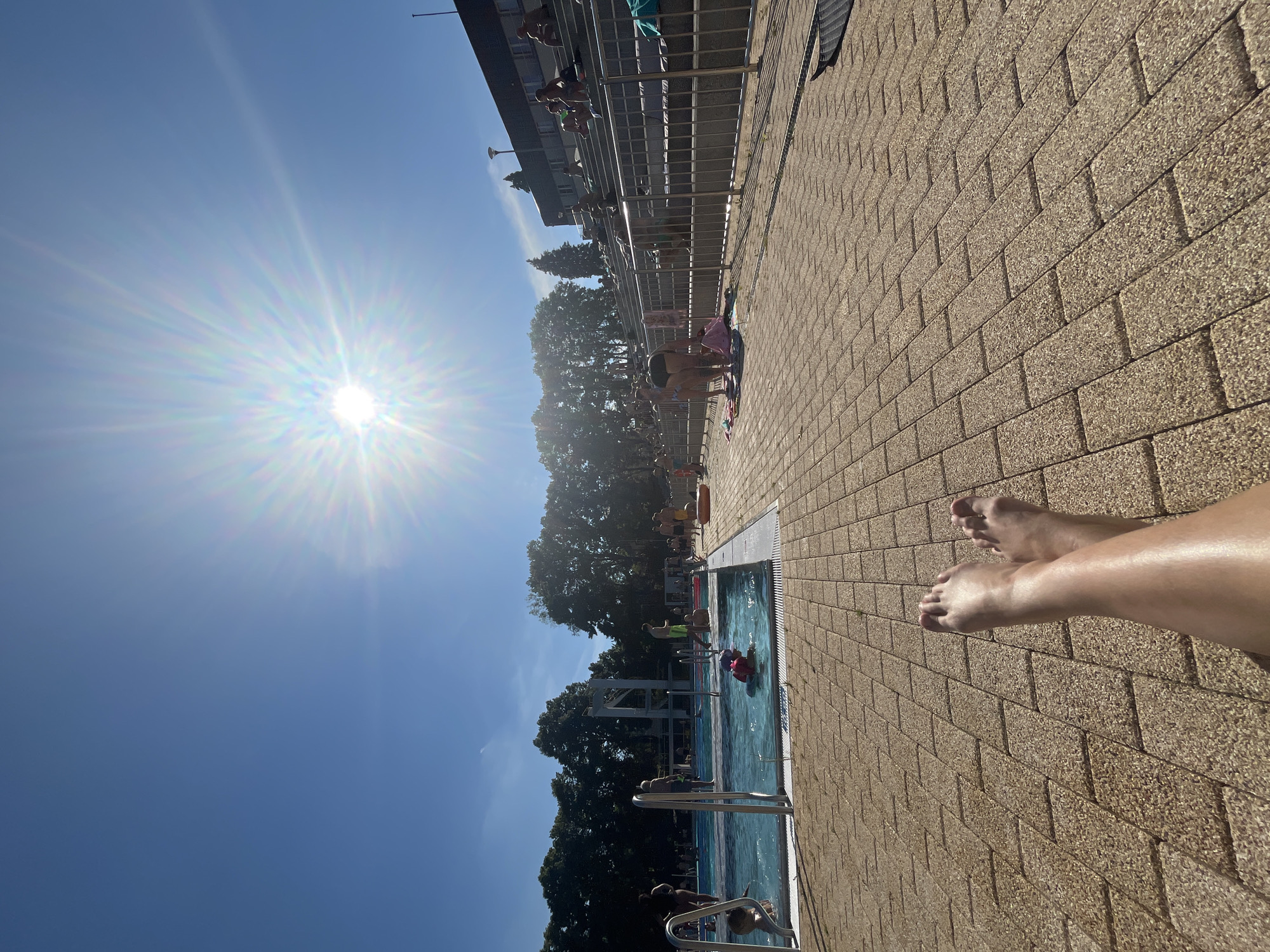 The image depicts a sunny day at an outdoor swimming pool. The sun is bright and high in the sky, casting a lens flare across the scene, suggesting it is midday. The weather appears to be warm and clear, with a vibrant blue sky.
The setting is a poolside area paved with light brown bricks. On the right side of the image, a person's bare feet and legs are visible, resting on the brickwork, implying they are relaxing or sunbathing. The skin appears tanned.
In the background, a swimming pool is filled with clear blue water. Several people are in and around the pool, including children playing in the water and adults sitting on the edge. There's a diving board structure visible, indicating a recreational facility. Trees and a building can be seen beyond the pool area. The overall mood seems relaxed and summery.
The orientation of the image is such that the sky and sun are on the left, and the poolside and pool are on the right. The brickwork extends diagonally into the distance. There is no visible text in the image.