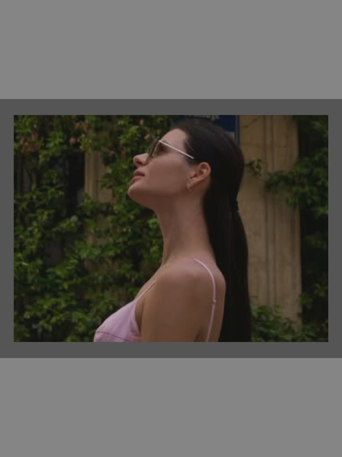 An outdoor medium shot captures a young woman from the chest up, facing left and looking upwards. She has dark hair and is wearing sunglasses and a light pink camisole. The background features lush green foliage and a textured stone pillar, suggesting an urban or garden setting.