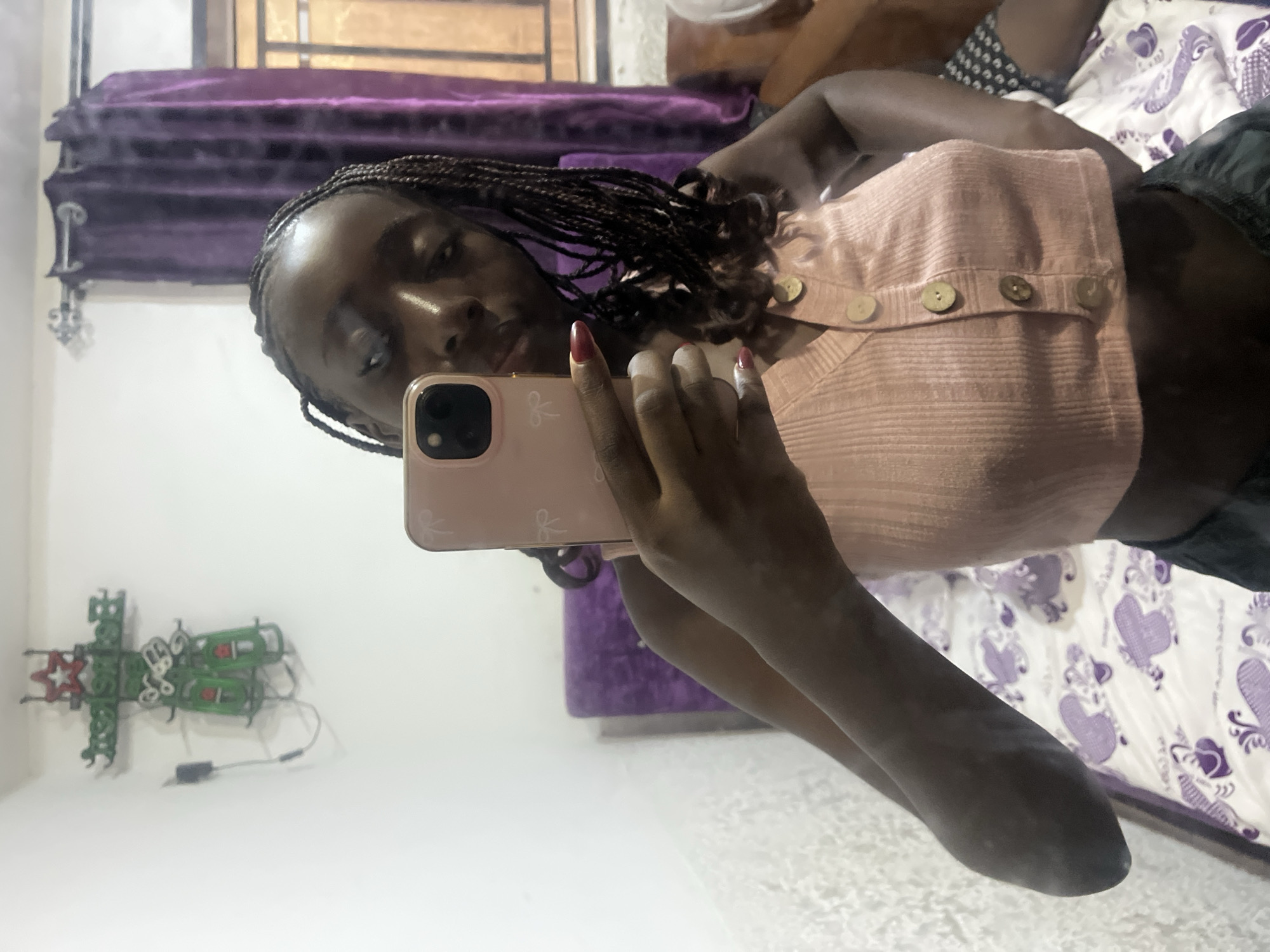 The image depicts a young woman with braided hair taking a selfie using a pink smartphone. She is wearing a light pink button-down top. The background includes a bed with a white and purple heart-patterned bedspread, purple curtains, and a wall. The location appears to be an interior space, potentially a bedroom, within the city of Abuja, Nigeria. There is a green neon-like lighting fixture on the wall.