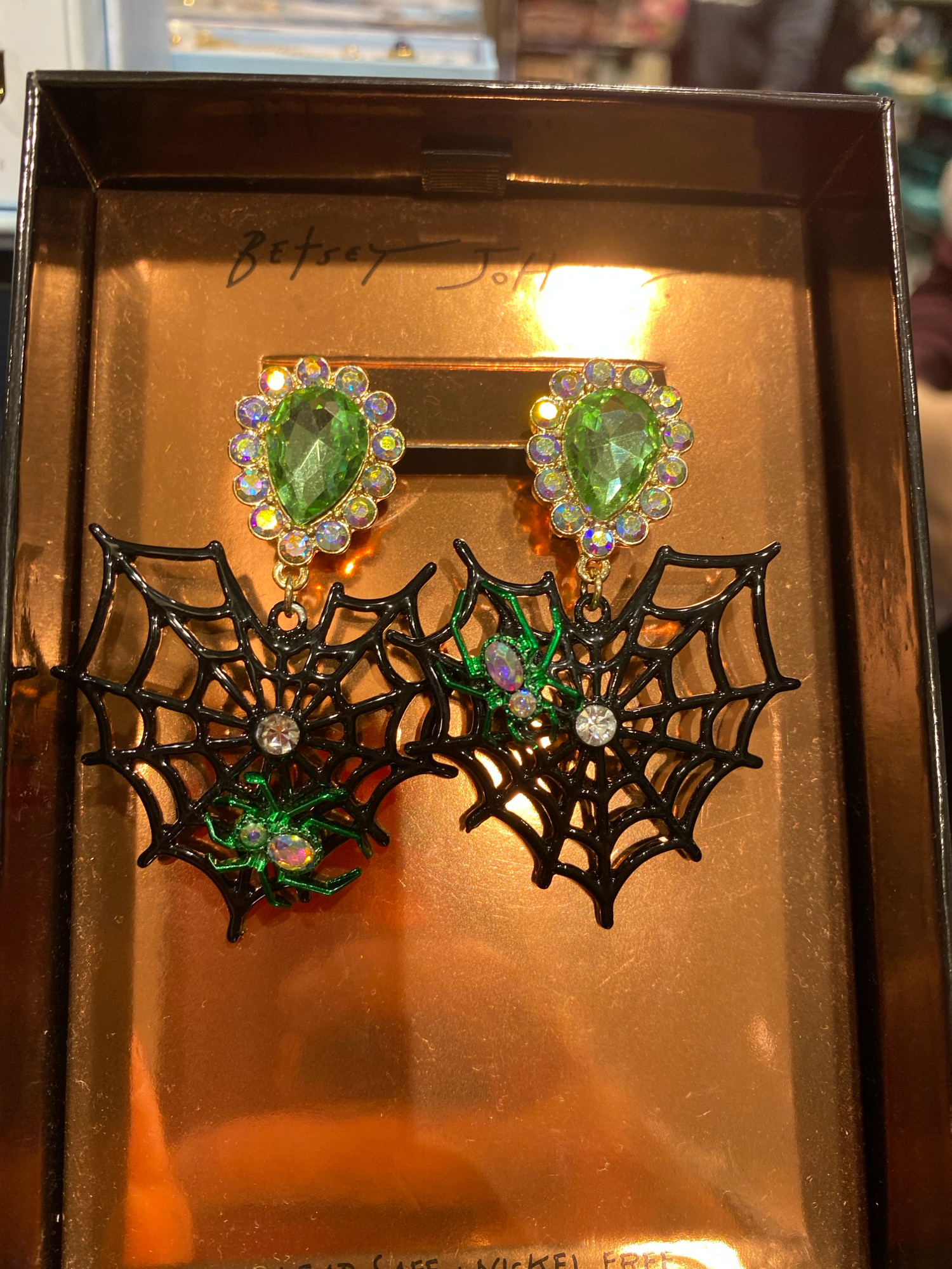 The image shows a pair of Halloween-themed earrings inside a gift box. The earrings are designed as black spiderwebs with green crystal spiders and a large green gemstone at the top, surrounded by smaller iridescent crystals. The gift box has a metallic copper interior and a black lid with the brand name "Betsey Johnson" handwritten on it. The background is blurred, suggesting an indoor setting, possibly a retail store. There are no people or specific architectural features visible that would allow for determining a location in Vienna, Austria.