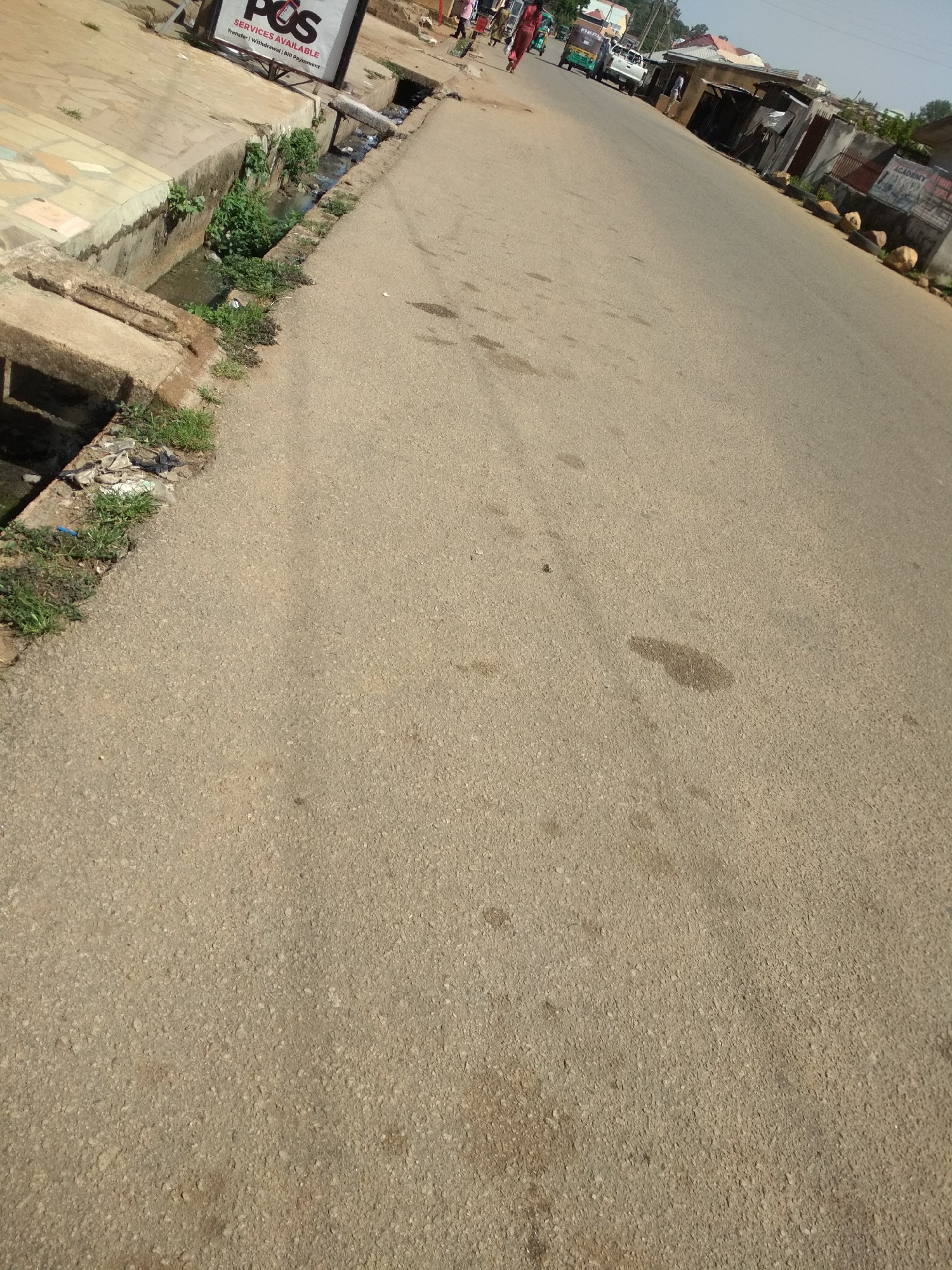 The image captures an outdoor scene along a paved road in what appears to be a busy, possibly semi-urban or commercial area, under bright daylight. The road, made of light-colored asphalt, stretches diagonally from the foreground into the distance, showing some dark stains and general wear.
On the left side of the road, a concrete drainage ditch or gutter runs parallel to the street. It contains stagnant, murky water and is heavily overgrown with green vegetation and weeds. Some scattered trash and debris are visible along the edge of the gutter and near the sidewalk. A concrete sidewalk with some patterned slabs is partially visible adjacent to the gutter. Above the gutter, a white sign with black text is clearly visible, reading: "POS", "SERVICES AVAILABLE", and "Transfer | Withdrawal | Bill Payment".
In the background, on the left side of the road, several people are walking. One person in a red outfit walks away from the camera. A light-colored pickup truck and a green and yellow tricycle (possibly an auto-rickshaw) are seen on the road further back.
On the right side of the road, various simple buildings and structures line the street. These appear to be shops or residential units, some with corrugated metal roofs. Several large rocks are placed along the roadside. A partially obscured sign on one of these buildings in the distance shows the word "ACADEMY".
The time of day appears to be mid-day, indicated by the strong, clear sunlight and bright conditions. The weather is sunny with a clear sky visible at the top right. No particular event is taking place, but rather everyday activity is depicted with people moving along the street. The overall impression is one of a functional, lived-in environment.