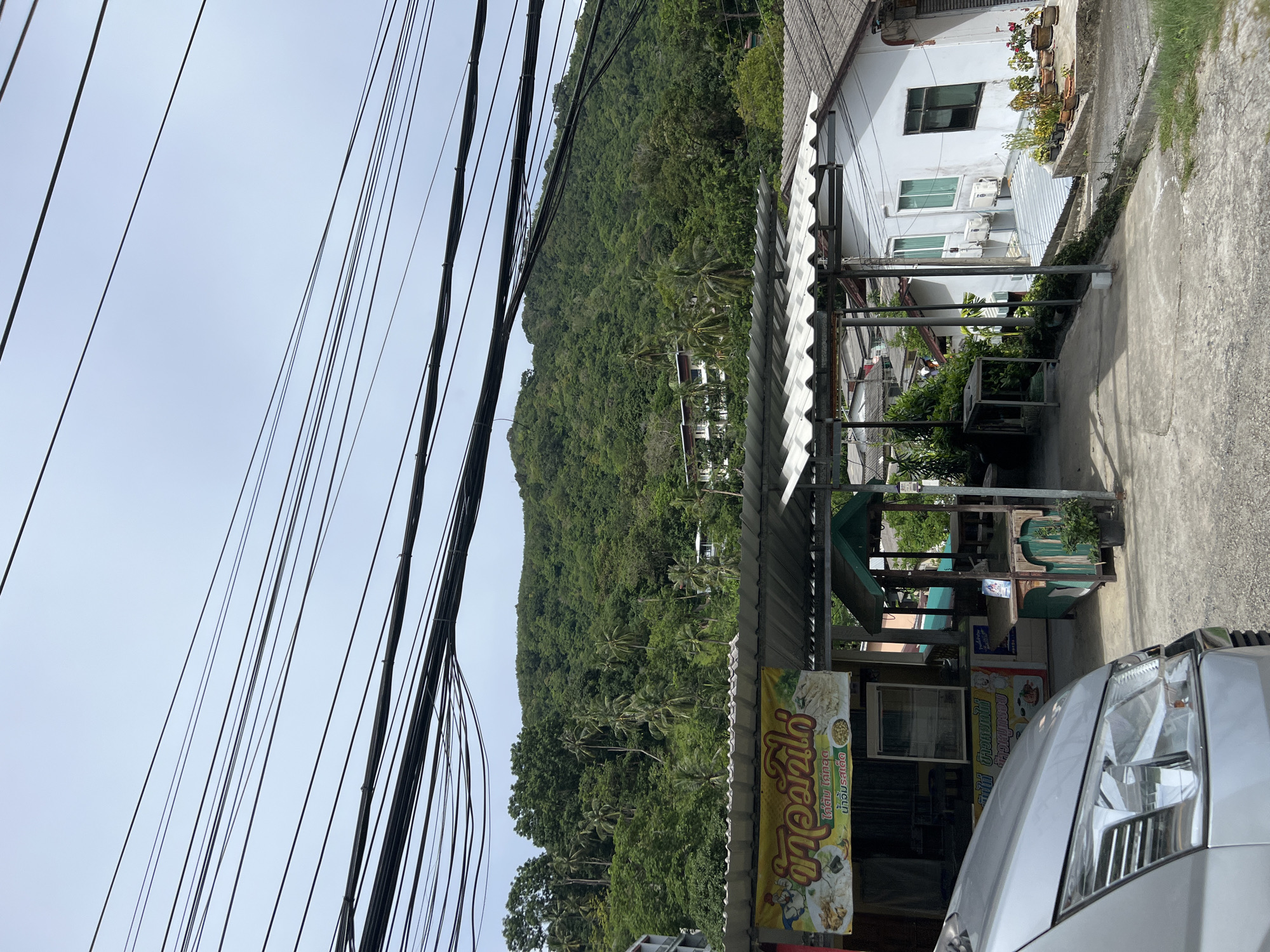 The image depicts a street scene in Karon, Thailand, with a prominent hillside covered in lush green vegetation dominating the background. In the foreground, a silver vehicle's headlight is visible on the right side. A small, open-air establishment with a corrugated metal roof is situated in the mid-ground, featuring a yellow banner with Thai text. This banner appears to advertise food with images of dishes like noodles and seafood. Shelving units and display cases are visible within the establishment, along with some potted plants. Above the street, a dense network of black electrical wires stretches across the sky, creating a strong diagonal line against the overcast sky. A white building with multiple windows and air conditioning units stands adjacent to the street and the establishment. The overall impression is a typical street view in a tropical, possibly coastal, area. The time of day appears to be daytime, with diffused light suggesting an overcast or hazy sky. There are no people clearly visible in the image.
