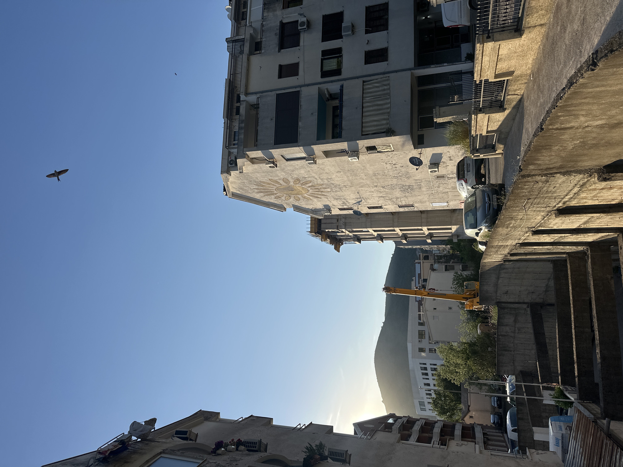 This eye-level, medium shot shows a street in Budva, Montenegro. The photograph looks upward at a steep angle. The sky is a clear, bright blue with two small birds flying in the upper left quadrant. Buildings flank both sides of the image. The left side shows older, multi-story buildings with weathered facades. One building has laundry drying on a rack outside a window. The right side features a modern, concrete construction, with exposed beams and a concrete retaining wall. In the center, a yellow construction crane is visible, with a mountain in the background and sunlight shining through the gap between buildings, creating a hazy effect. Several cars are parked in the street.