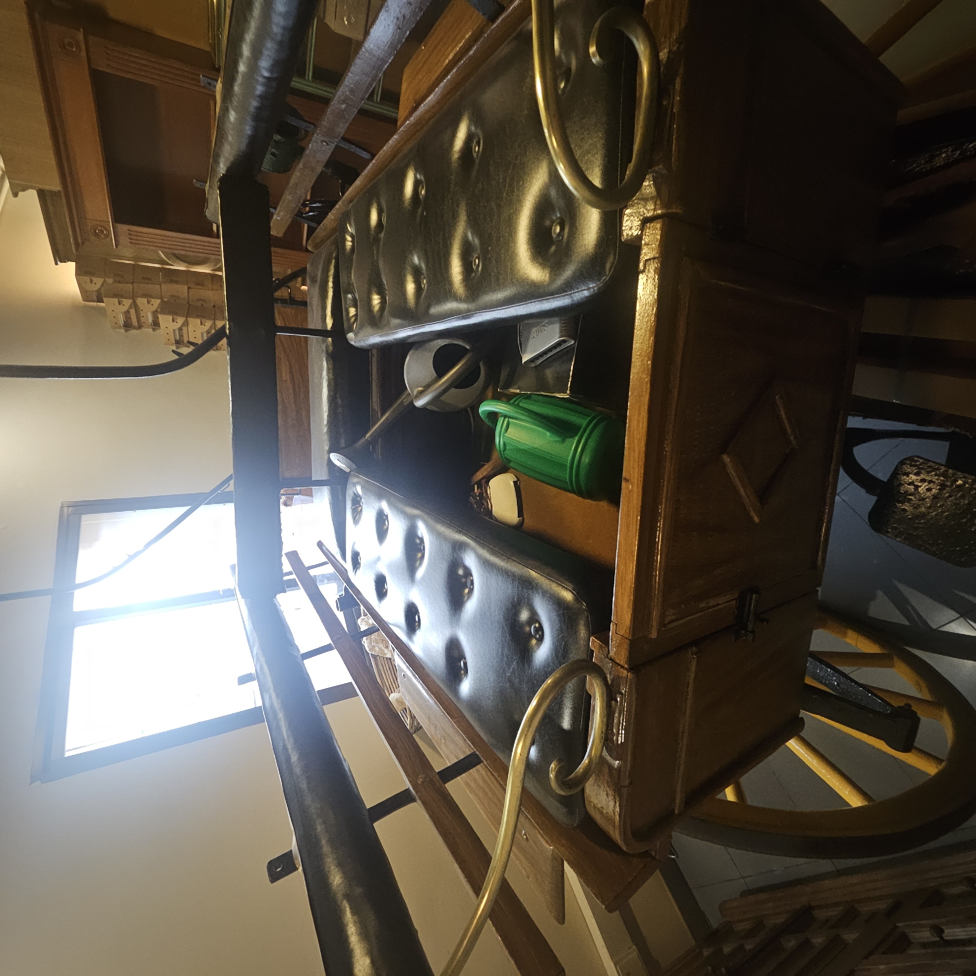 The image shows an interior scene, possibly a shop or storage area, in Almadrones, Spain. The primary object is a wooden carriage with black cushioned seats. The carriage has large yellow spoked wheels. Inside the carriage, there is a green watering can and other indistinct objects. In the background, there is a large wooden piece of furniture, possibly a cabinet or fireplace surround, and stacks of cardboard boxes. A large window with bright light coming through it is visible on the left side of the image. The scene appears to be lit by natural light from the window and possibly some artificial lighting. There are no people visible in the image.
