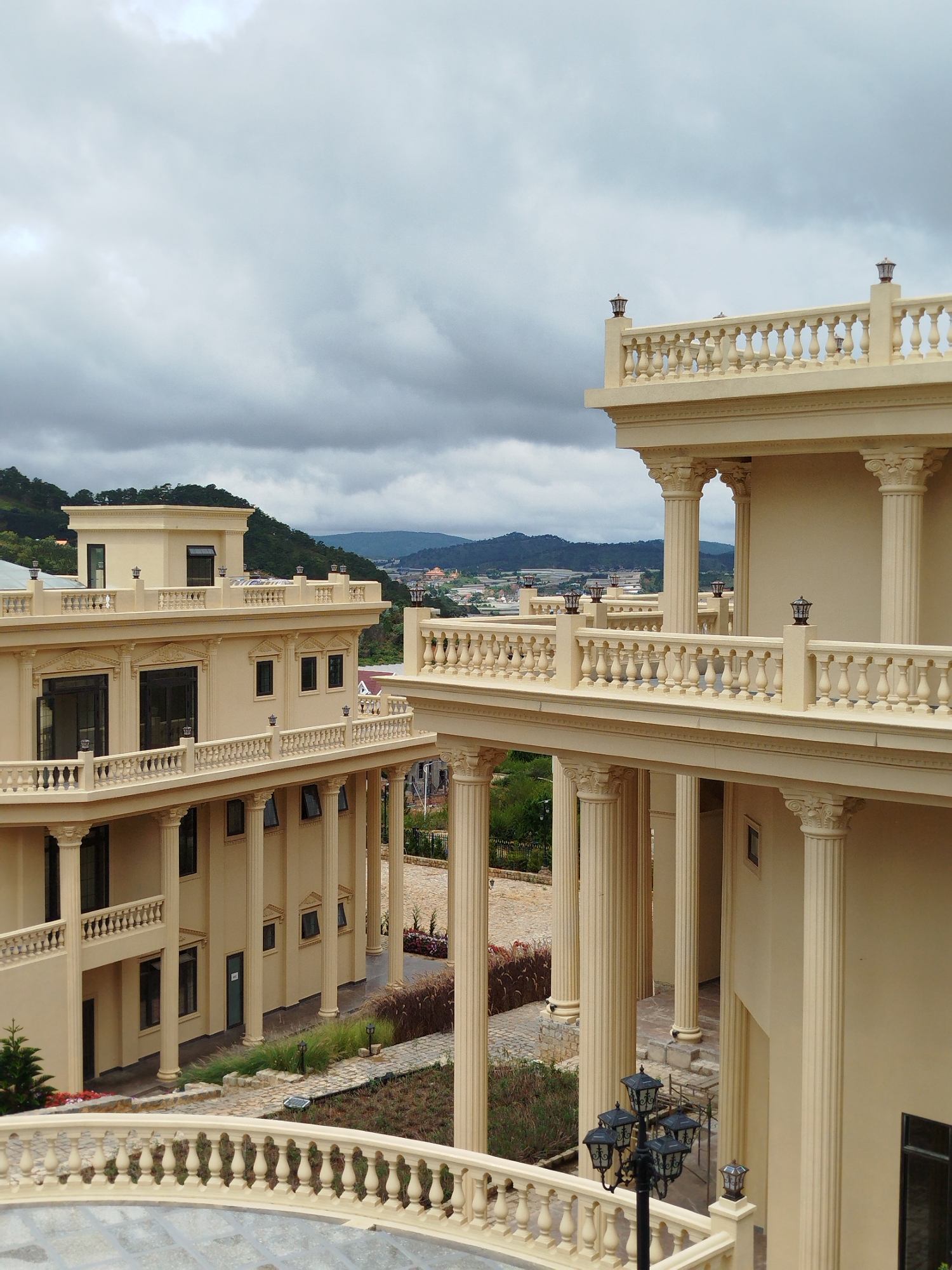 The image shows a building with classical architectural elements, including columns and ornate railings, likely in Lạc Dương, Vietnam. The building is pale yellow and has multiple stories with balconies and large windows. The exterior is decorated with decorative moldings and lamps. In the foreground, a curved railing and a multi-headed lamppost are visible. The background features a hilly landscape with lush green vegetation and other buildings in the distance, under a cloudy sky. The scene appears to be a resort or hotel due to its grand architecture and scenic location. There are no people visible in the image.