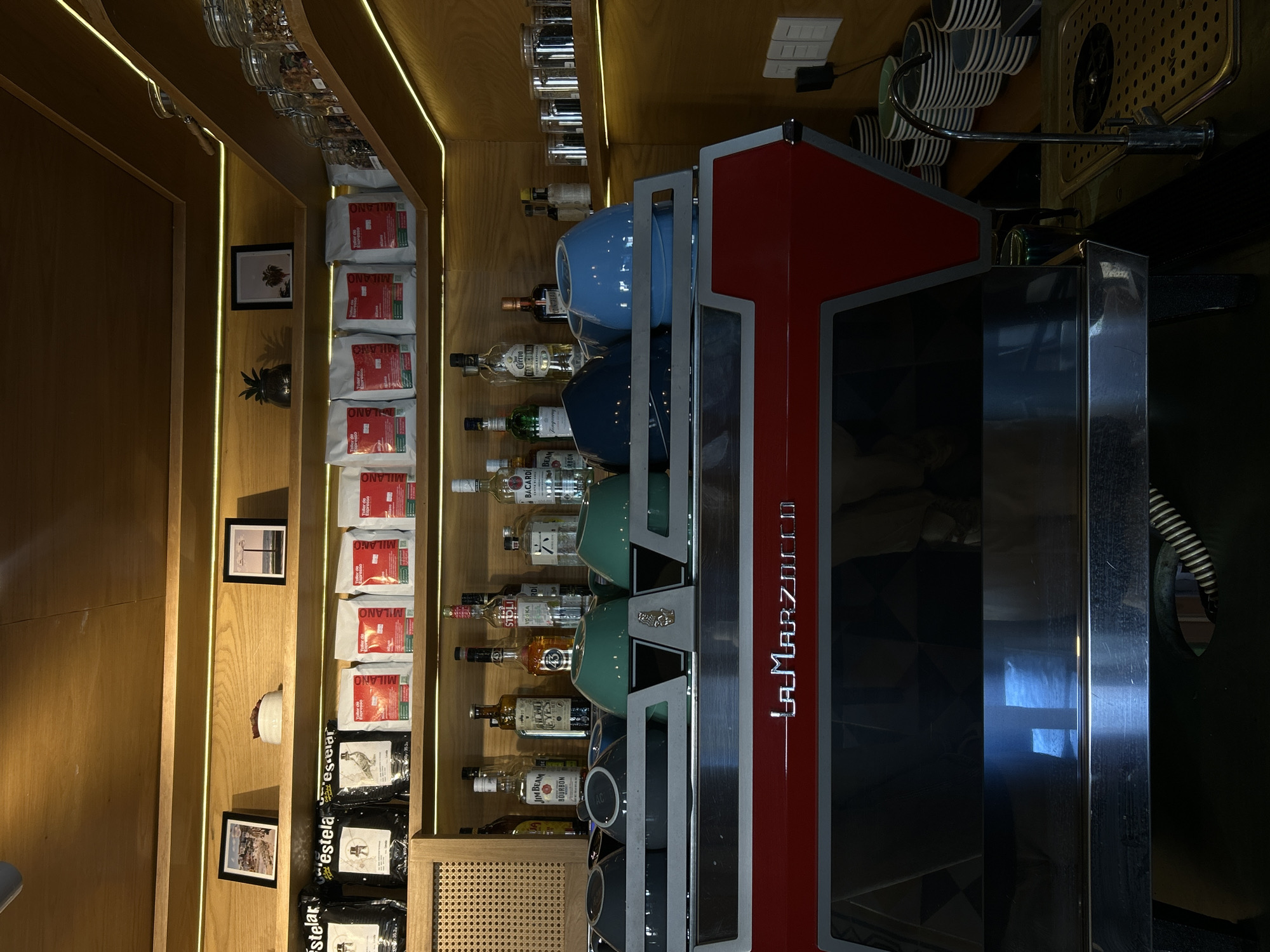 The image shows the interior of a bar or cafe, potentially in Mazatlán, Mexico, given the 'Estelar' branding visible in the lower left. A La Marzocco espresso machine, with stacked mugs in varying colors (blue, green, gray), dominates the right side of the frame. Bottles of alcohol, including brands such as Bacardi, Stolichnaya, Jim Beam, and Jose Cuervo, are displayed on shelves behind the machine. The left side of the image features a wooden shelf with bags of coffee labeled "Milano" and "Estelar" and framed pictures are placed on the wall. Glass jars are positioned on the top shelf. The lighting is a combination of natural and artificial, with LED strips running along the shelf edges. The scene appears to be well-stocked and organized for commercial use.