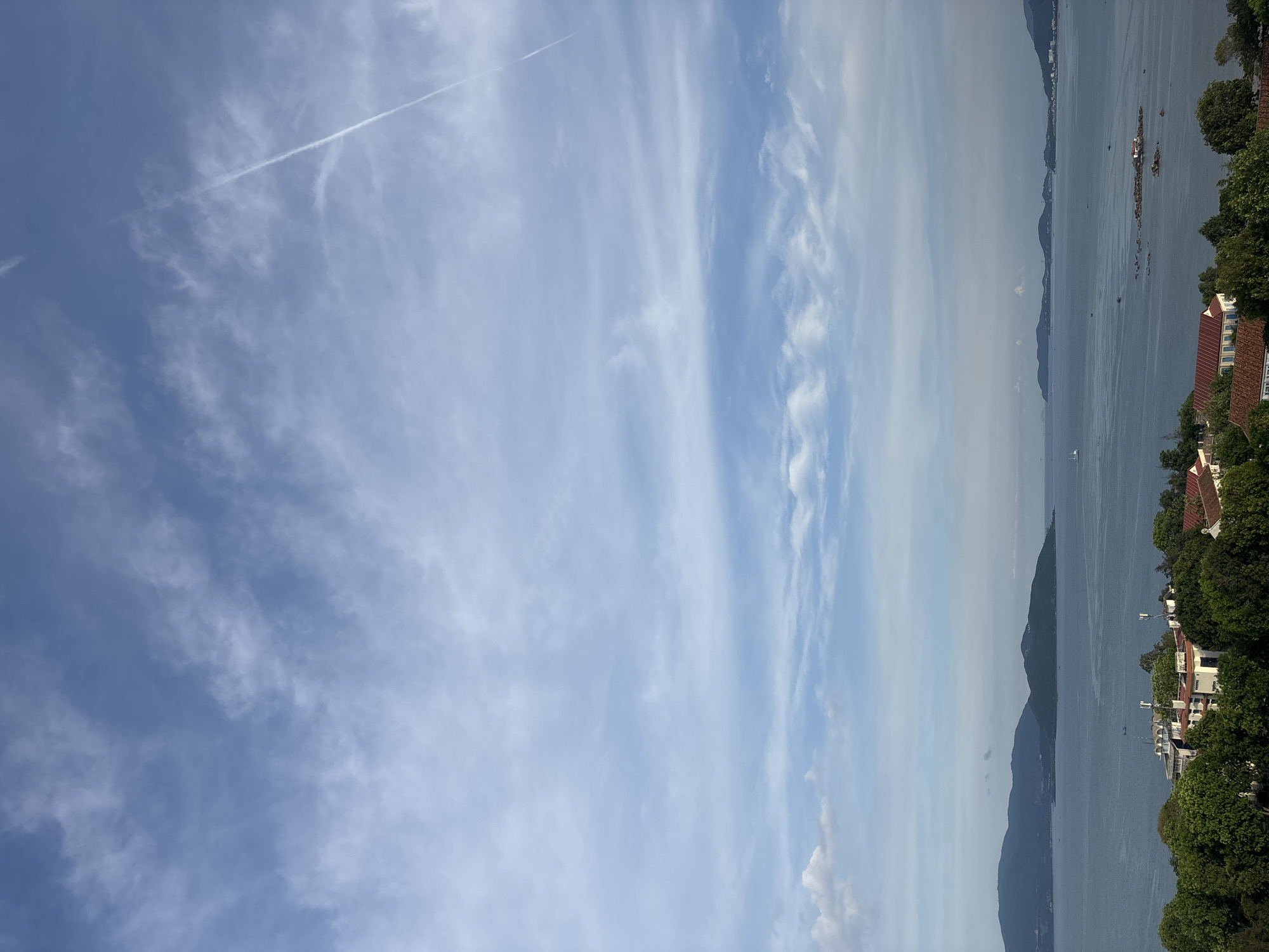 The image depicts a scenic view of a calm body of water, likely a bay or sea, with several islands and coastal landscapes in the distance. The sky is a pale blue with scattered wispy clouds, and a distinct contrail from an aircraft stretches diagonally across the upper right portion of the frame. In the foreground, there is a densely vegetated area with lush green trees, and nestled amongst them are several buildings with red-tiled roofs and white walls. Some of these structures appear to be residential or part of a resort. The water is a deep blue, reflecting the sky, and there are a few small boats visible on its surface, as well as some rocks protruding from the water near the coast. The overall atmosphere is serene and natural.