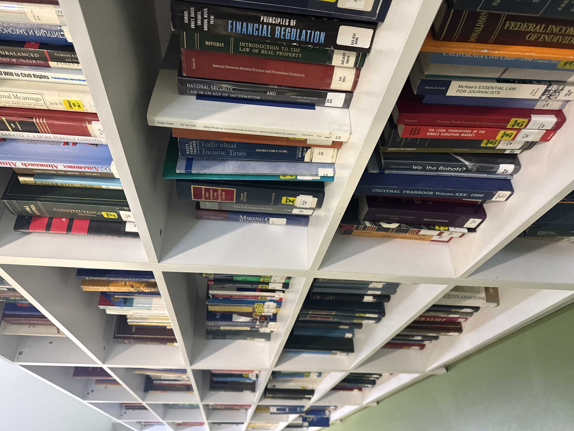 The image shows shelves in a library or legal office filled with books on various legal and financial topics. The books are organized in a white cube shelving unit. Some of the visible titles include "Principles of Financial Regulation," "National Security, Journalism, and Law in an Age of Information," "Individual Income Taxes," "Federal Income of Individuals," and "McNae's Essential Law for Journalists." The perspective is from below, looking up at the shelves, creating a sense of depth.